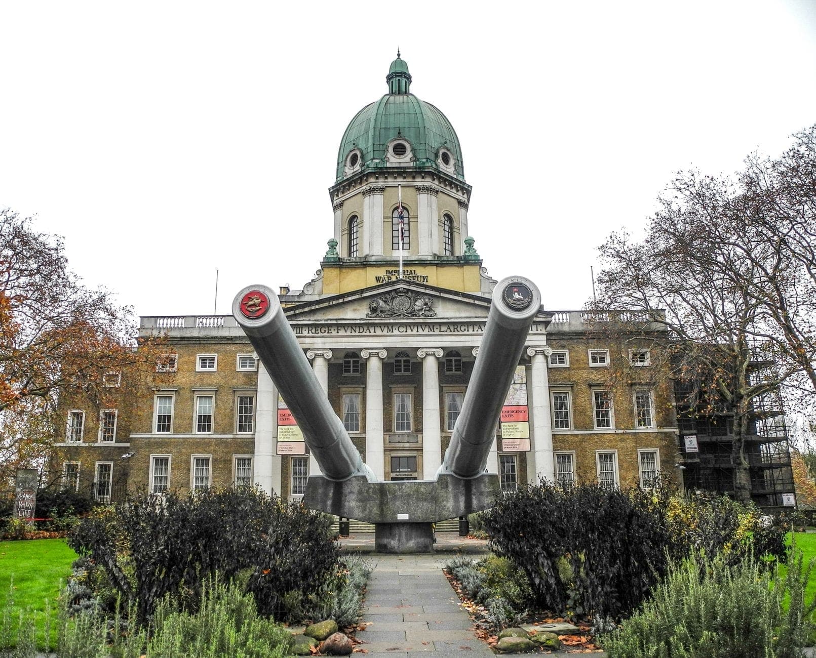 Widok z przodu na Imperialny Muzeum Wojenne w Londynie z charakterystyczną kopułą i wielkimi działami okrętowymi na pierwszym planie, otoczonymi drzewami i ogrodami.