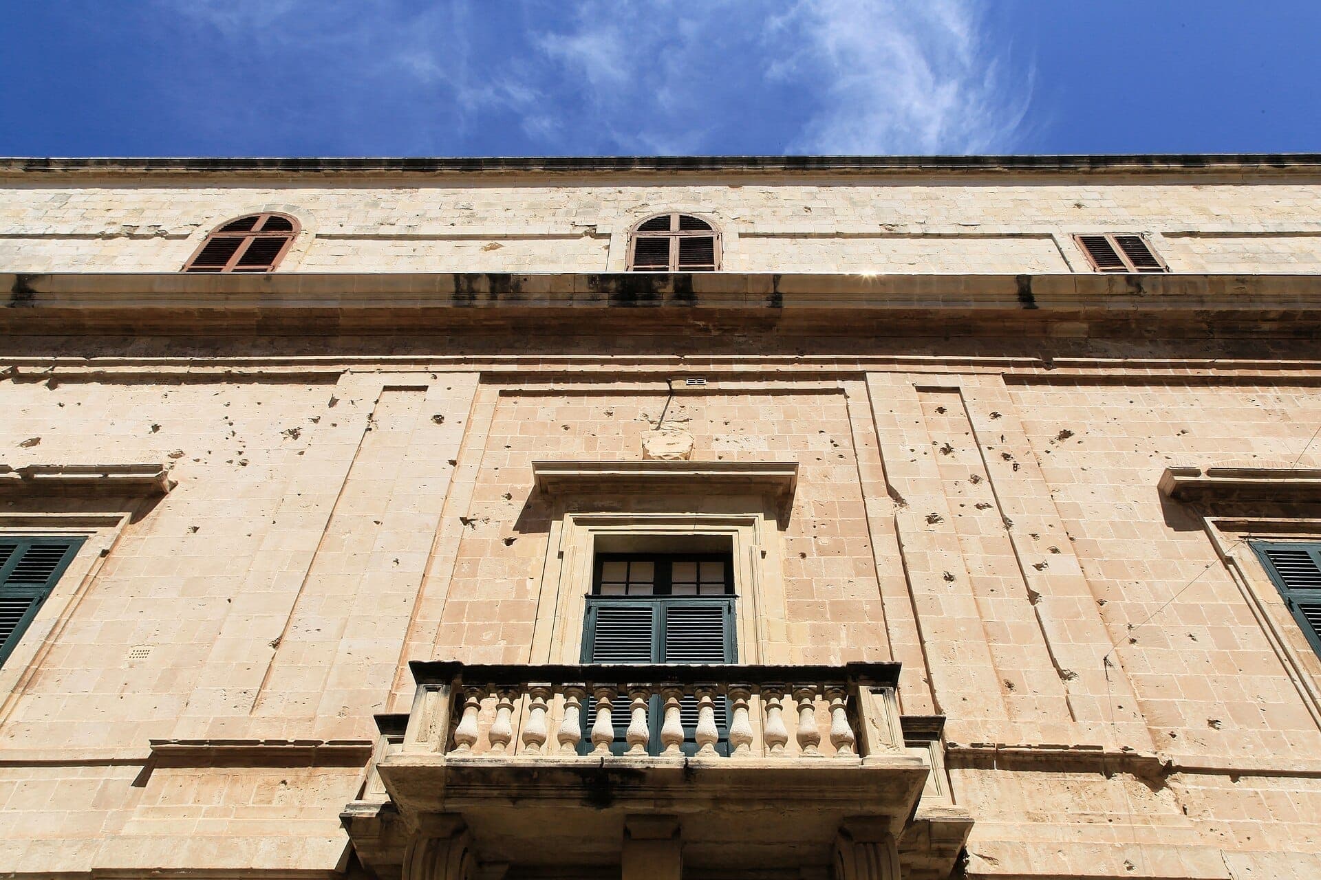 Fassade des Inquisitorenpalastes in Birgu mit verwitterten Kalksteinmauern, Fensterläden und einem Steinbalkon unter einem wolkenlosen blauen Himmel.