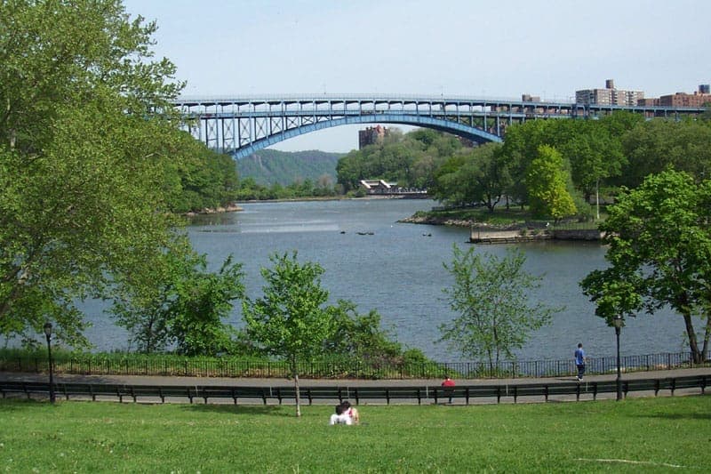 วิวของ Inwood Hill Park พร้อมต้นไม้เขียวขจี แม่น้ำ Harlem และสะพานโค้งเหล็กใต้ท้องฟ้าสีฟ้าใส