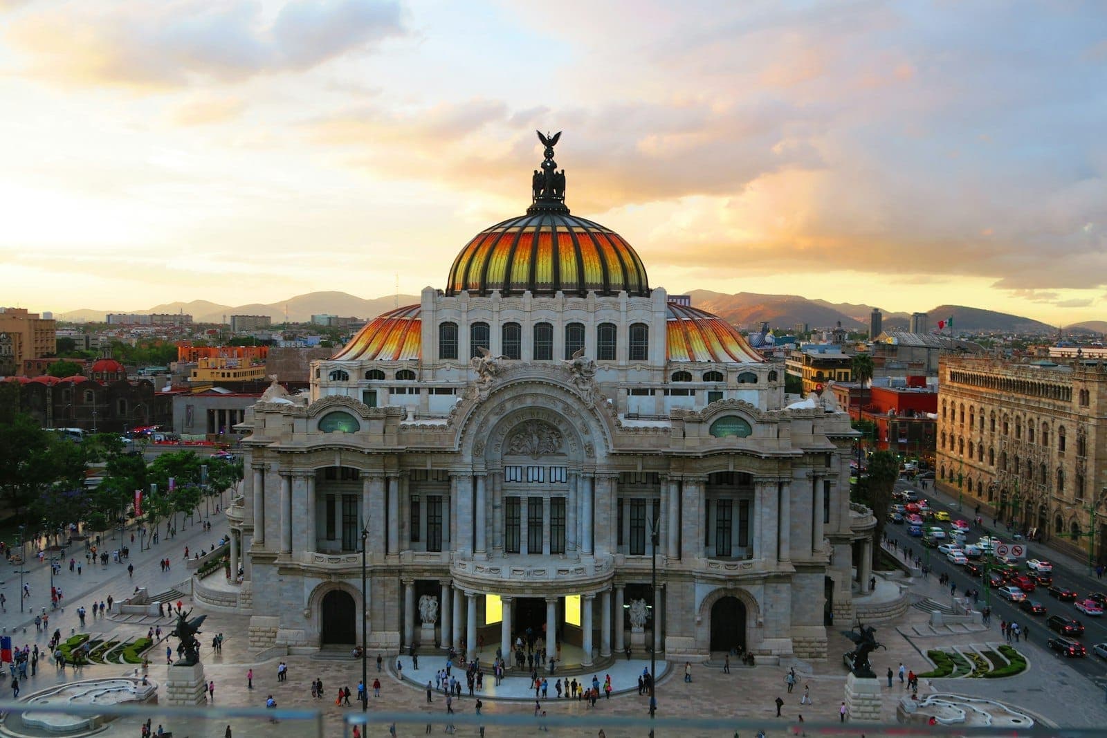 วิวกว้างของ Palacio de Bellas Artes ในเม็กซิโกซิตี้ช่วงพระอาทิตย์ตก เต็มไปด้วยผู้คน ตึกสูงล้อมรอบและมีภูเขาอยู่ฉากหลัง
