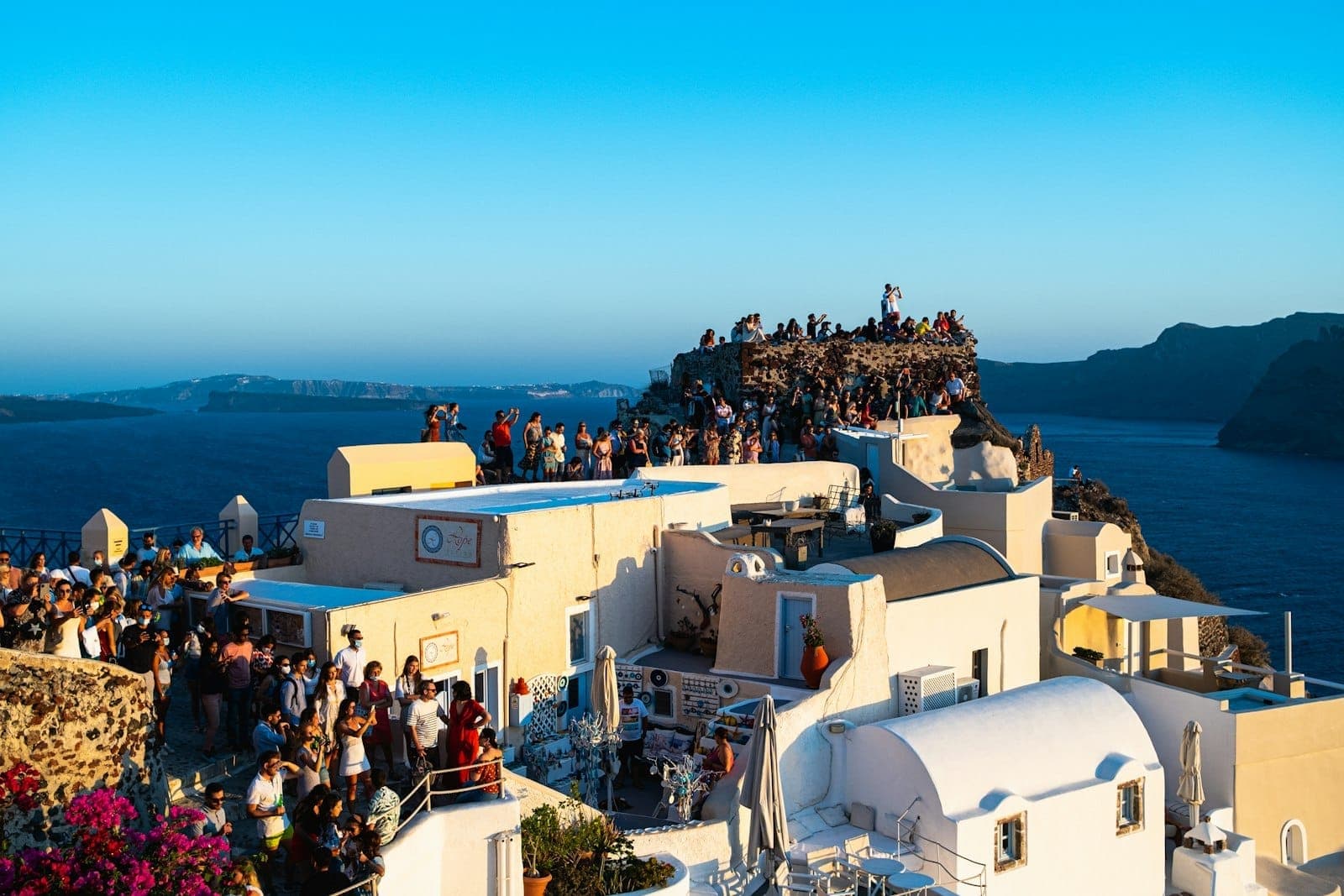 Große Menschenmengen auf den Dächern von Santorini und einem Aussichtspunkt bei Sonnenuntergang, die die belebte und überfüllte Touristenatmosphäre der Insel zeigen.
