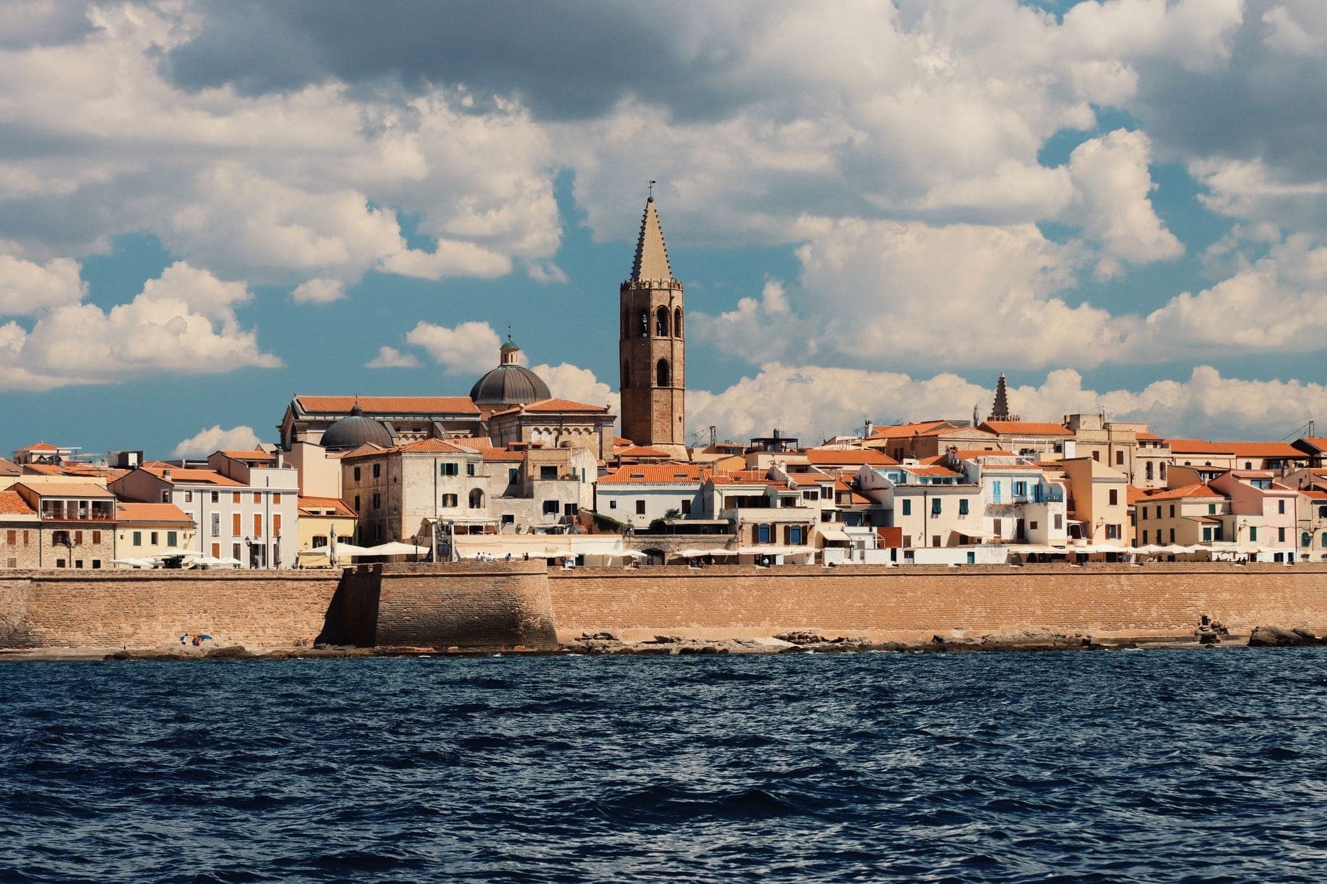 Localidad costera sarda con edificios de piedra y un alto campanario vistos desde el mar bajo un cielo con nubes dramáticas.