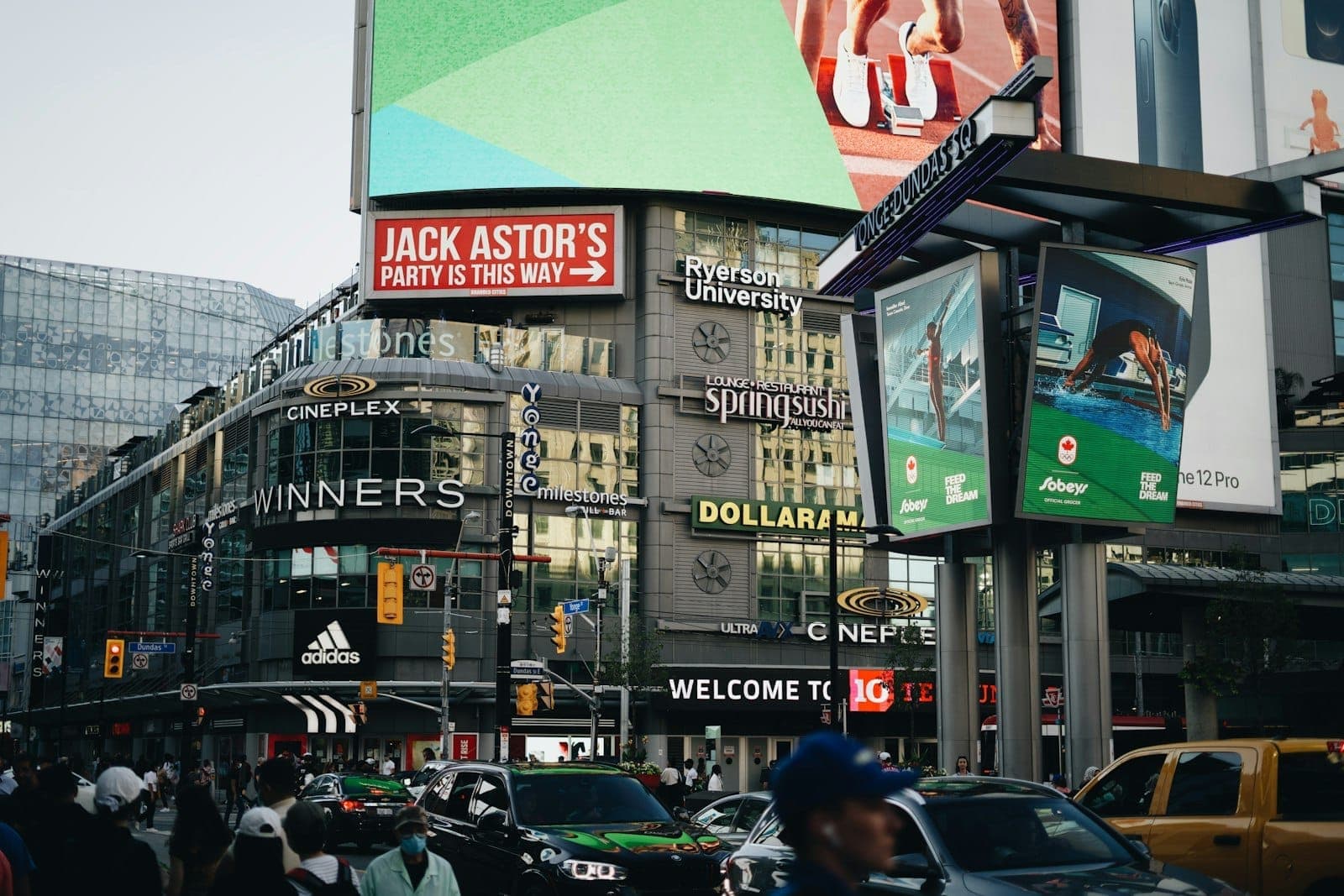 Busy downtown Toronto intersection at Yonge-Dundas Square, filled with people, cars, and recognizable city signage.