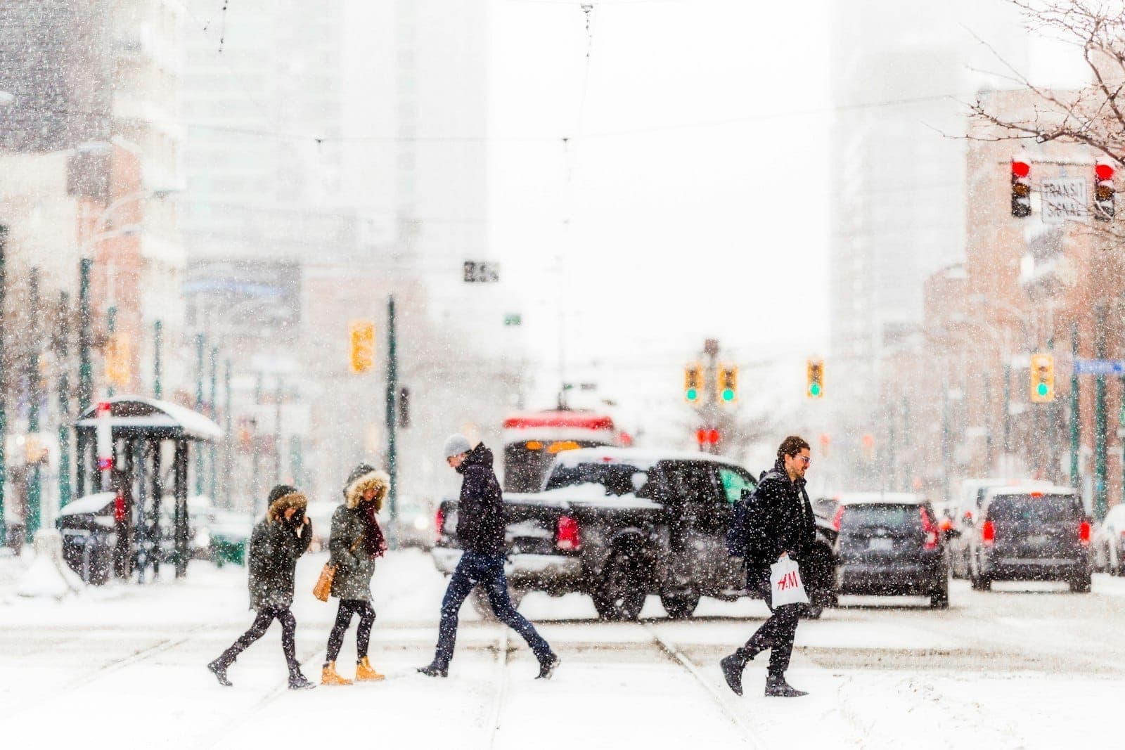 Pedestrians bundled up and crossing a busy Toronto street during heavy snowfall, with snow-covered roads and urban traffic lights visible.