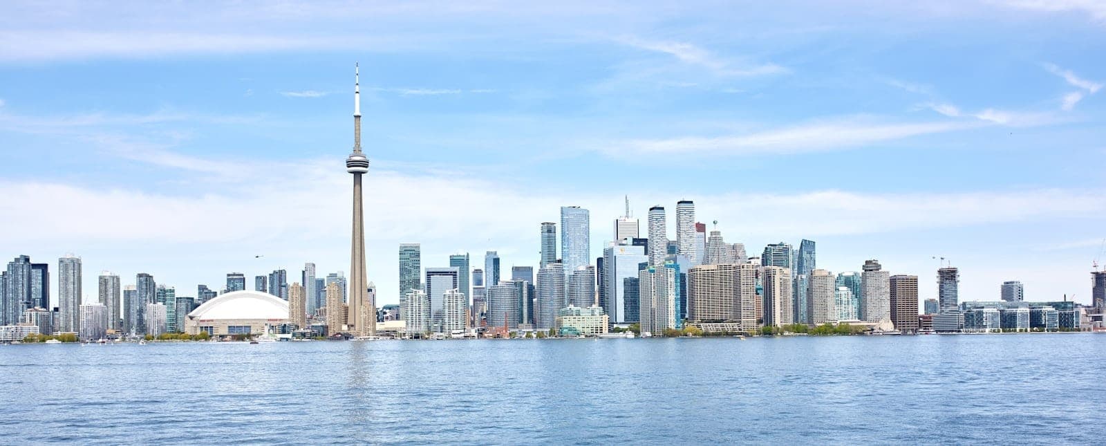 Panorama siang yang luas dan cerah dari cakrawala Toronto dengan CN Tower dan gedung-gedung pencakar langit, terlihat dari seberang air di bawah langit biru, memancarkan kesan aman dan energi kota yang ramah.