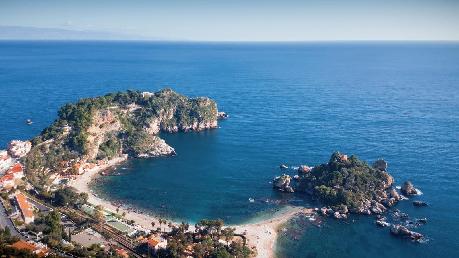 Tanaw mula sa itaas ng Isola Bella na nakakabit sa baybayin ng Taormina sa pamamagitan ng makitid na landas, napapalibutan ng turkesa at malinaw na Ionian Sea at luntiang burol.