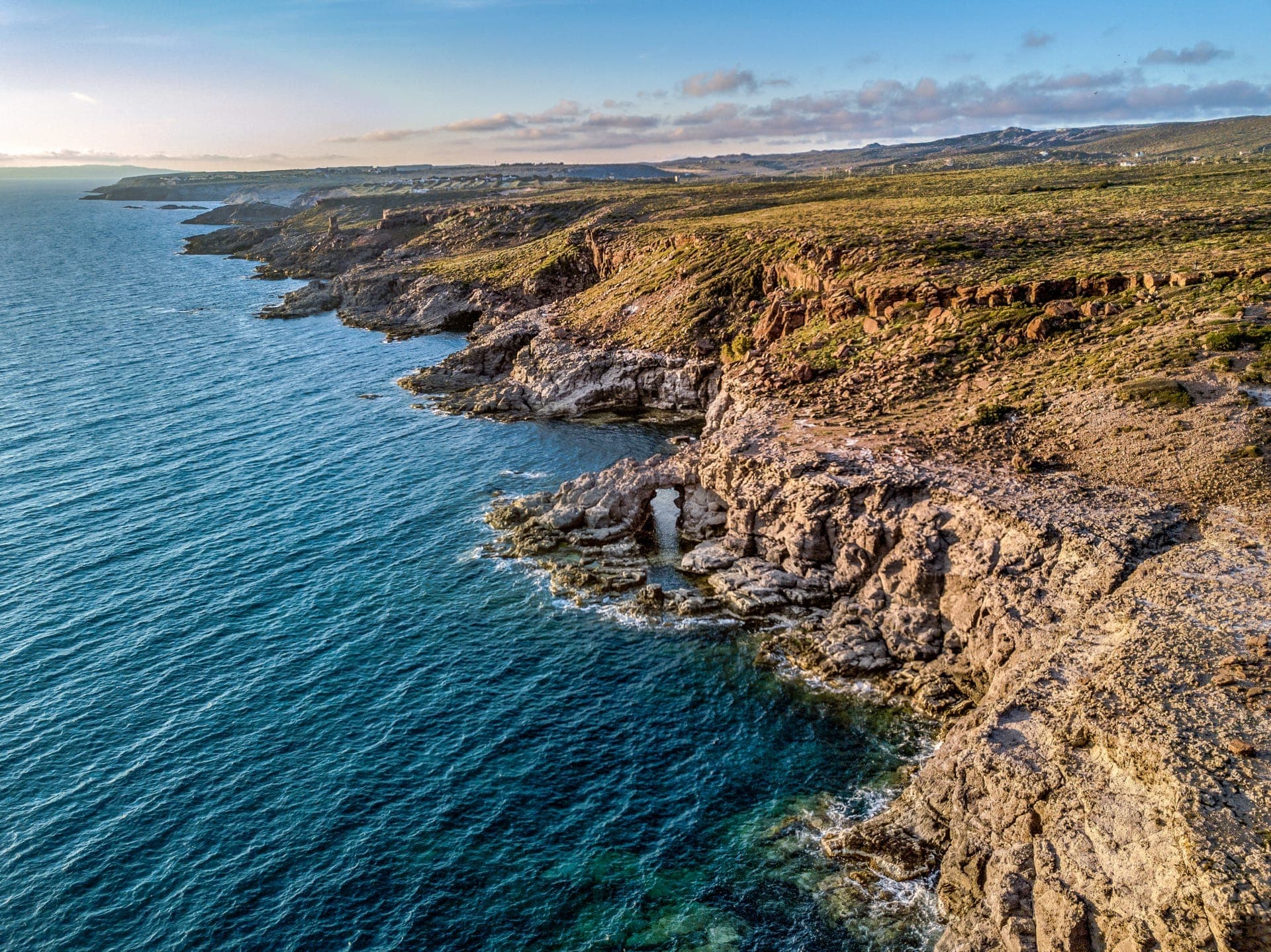 Costa rocosa de la Isola di Sant'Antioco con acantilados escarpados y mar azul cristalino bajo un cielo brillante en la hora dorada.