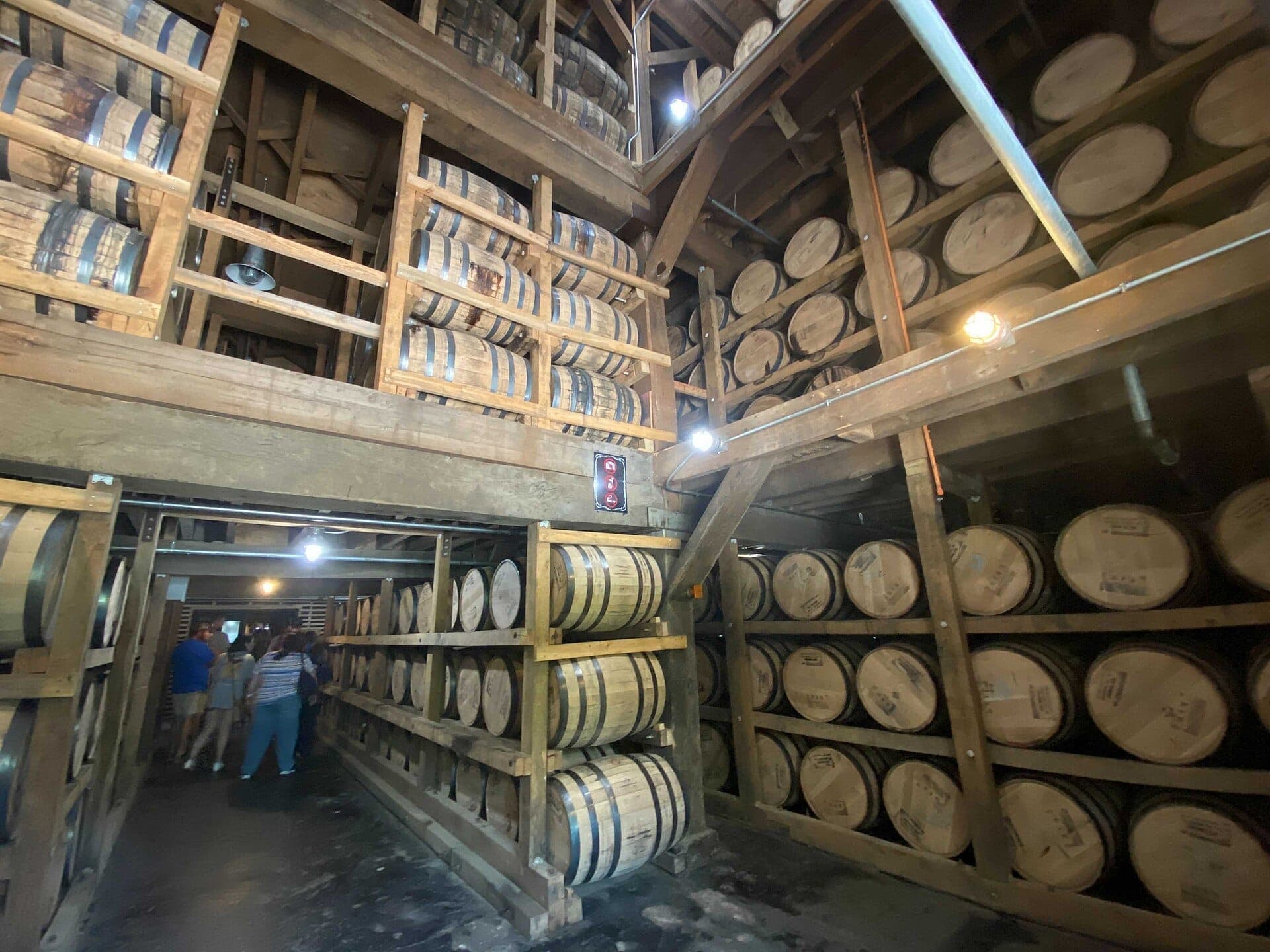 Interior view of Jack Daniel's Distillery barrel house, featuring rows of stacked whiskey barrels and a group of visitors on tour.