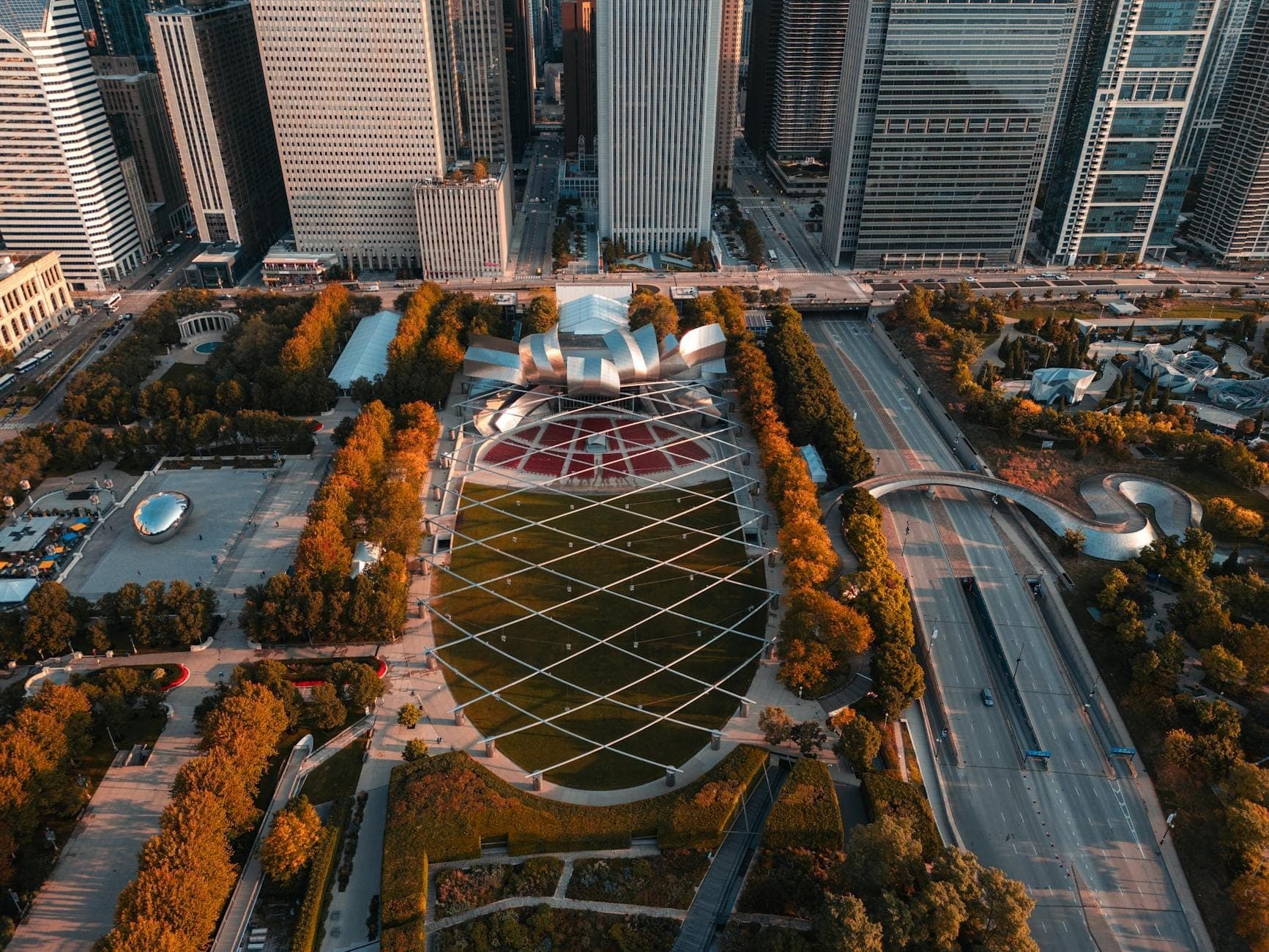 Luftaufnahme des Jay Pritzker Pavilion mit seiner markanten Metallkonstruktion, dem großen Rasen und den umliegenden Wolkenkratzern der Chicagoer Innenstadt an einem hellen Tag.