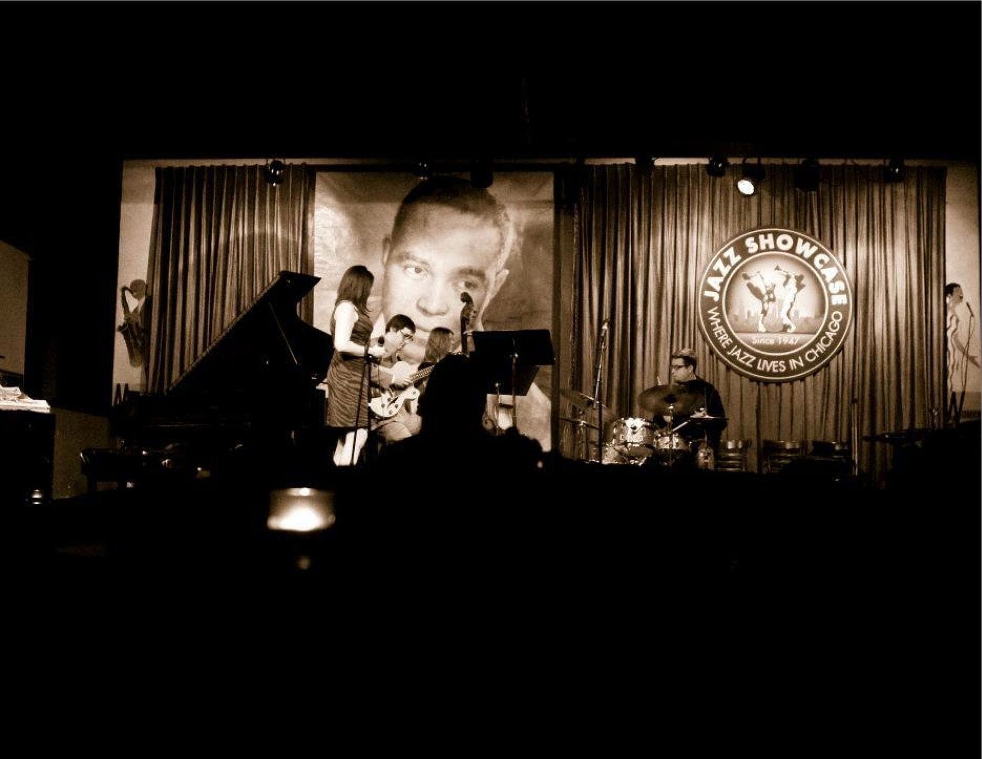 วงดนตรีแจ๊สสดกำลังแสดงบนเวทีที่แสงสลัวของ Jazz Showcase ในชิคาโก โดยมีนักดนตรีเล่นเปียโน กีตาร์ และกลอง