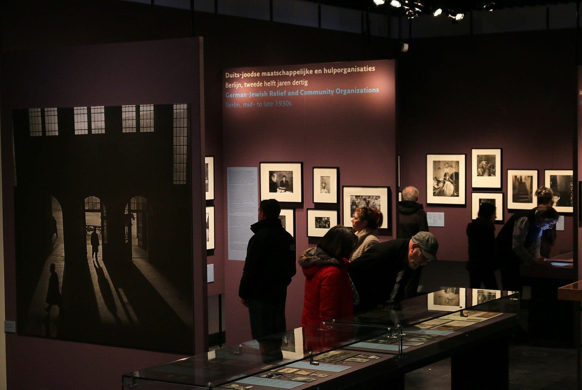 Besucher betrachten historische Fotografien und Artefakte in einer schwach beleuchteten Ausstellung im Jewish Museum Amsterdam, mit großen Schwarz-Weiß-Bildern an lila Wänden.