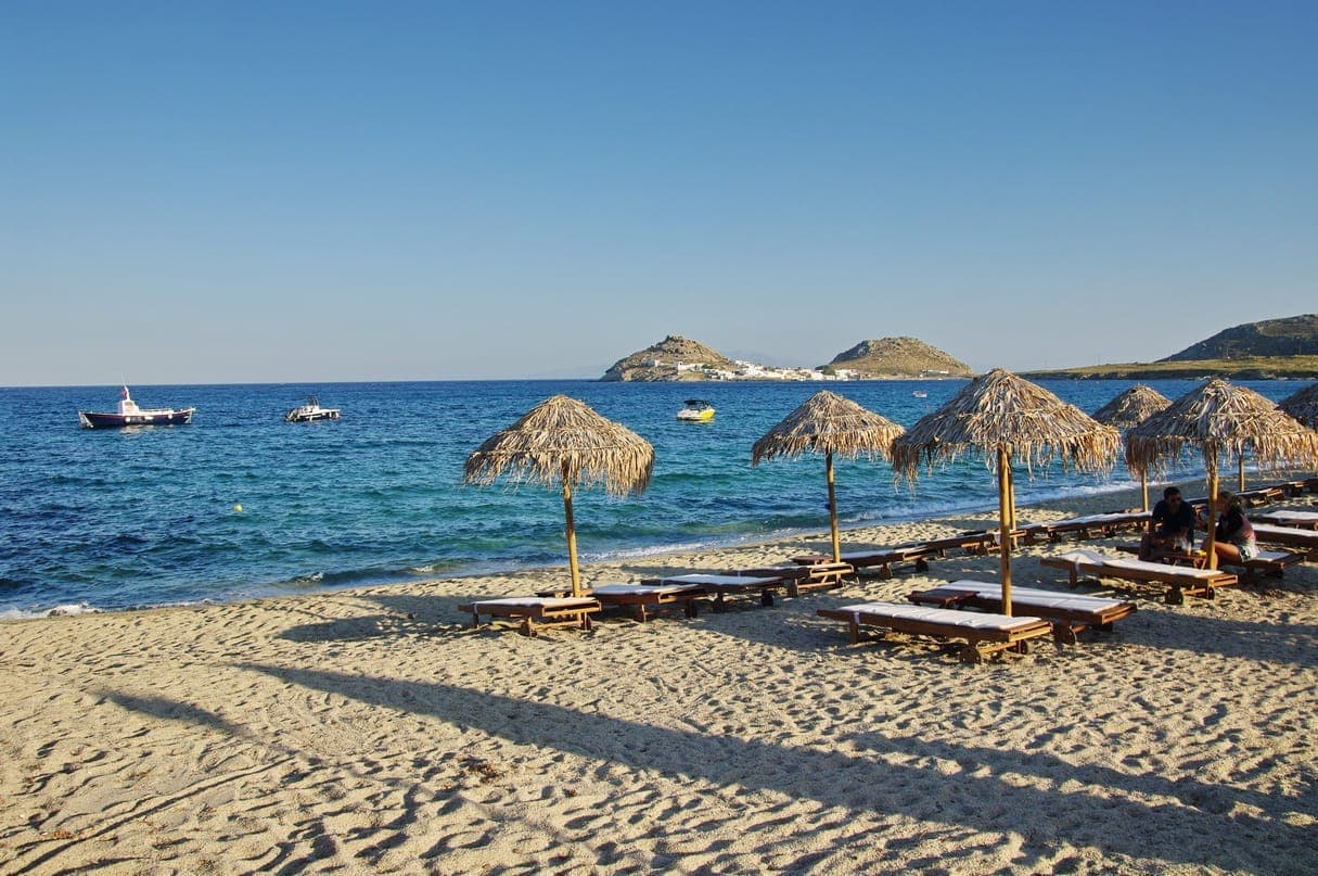 Sandiger Kalafatis Beach auf Mykonos mit Strohsonnenschirmen, Liegestühlen, klarem blauem Meer und fernen felsigen Hügeln unter einem sonnigen Himmel.