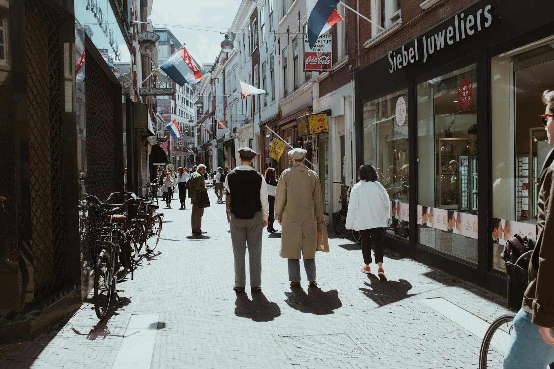 Fußgänger spazieren entlang der Kalverstraat in Amsterdam, mit auf dem Gehweg geparkten Fahrrädern, niederländischen Flaggen und Geschäften entlang der sonnenbeschienenen Straße.