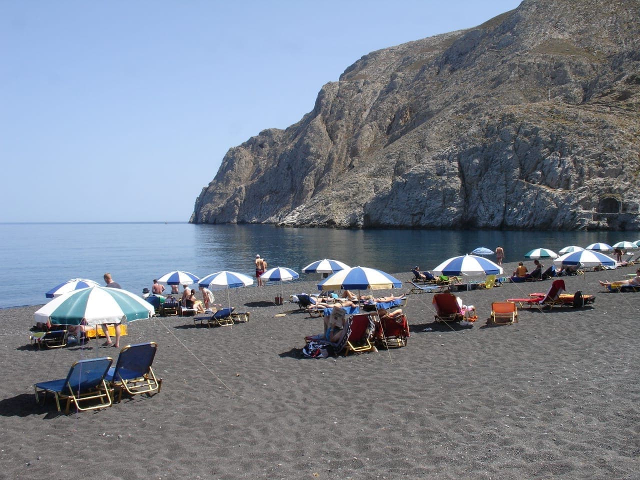 Kamari Beach auf Santorini mit Liegestühlen, Sonnenschirmen, entspannenden Menschen und beeindruckenden Felsklippen unter einem klaren blauen Himmel.