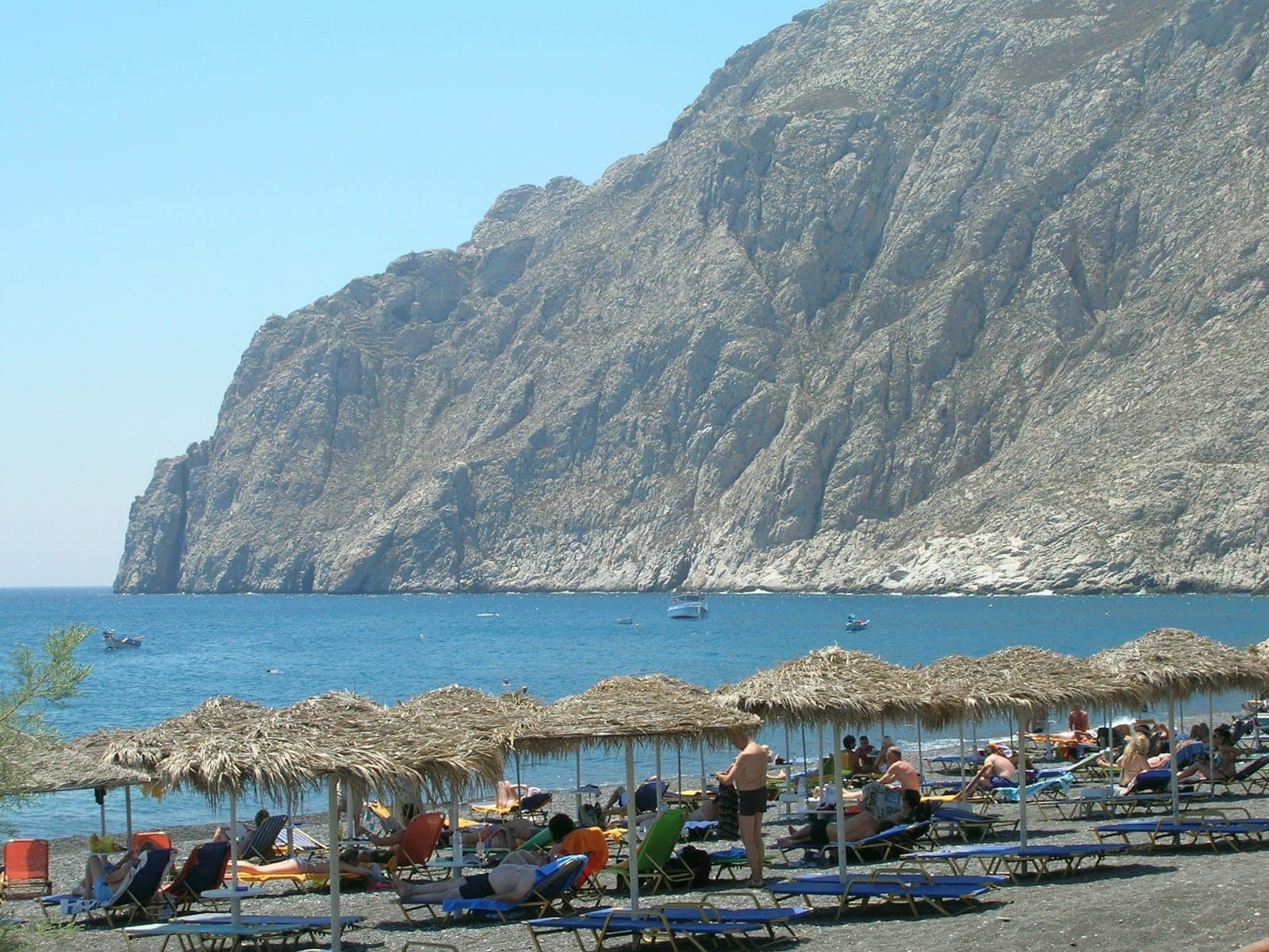 Sonnenanbeter entspannen sich am Schwarzkiesstrand von Kamari unter Strohschirmen, mit klarem blauem Meer und schroffen Felsklippen im Hintergrund.