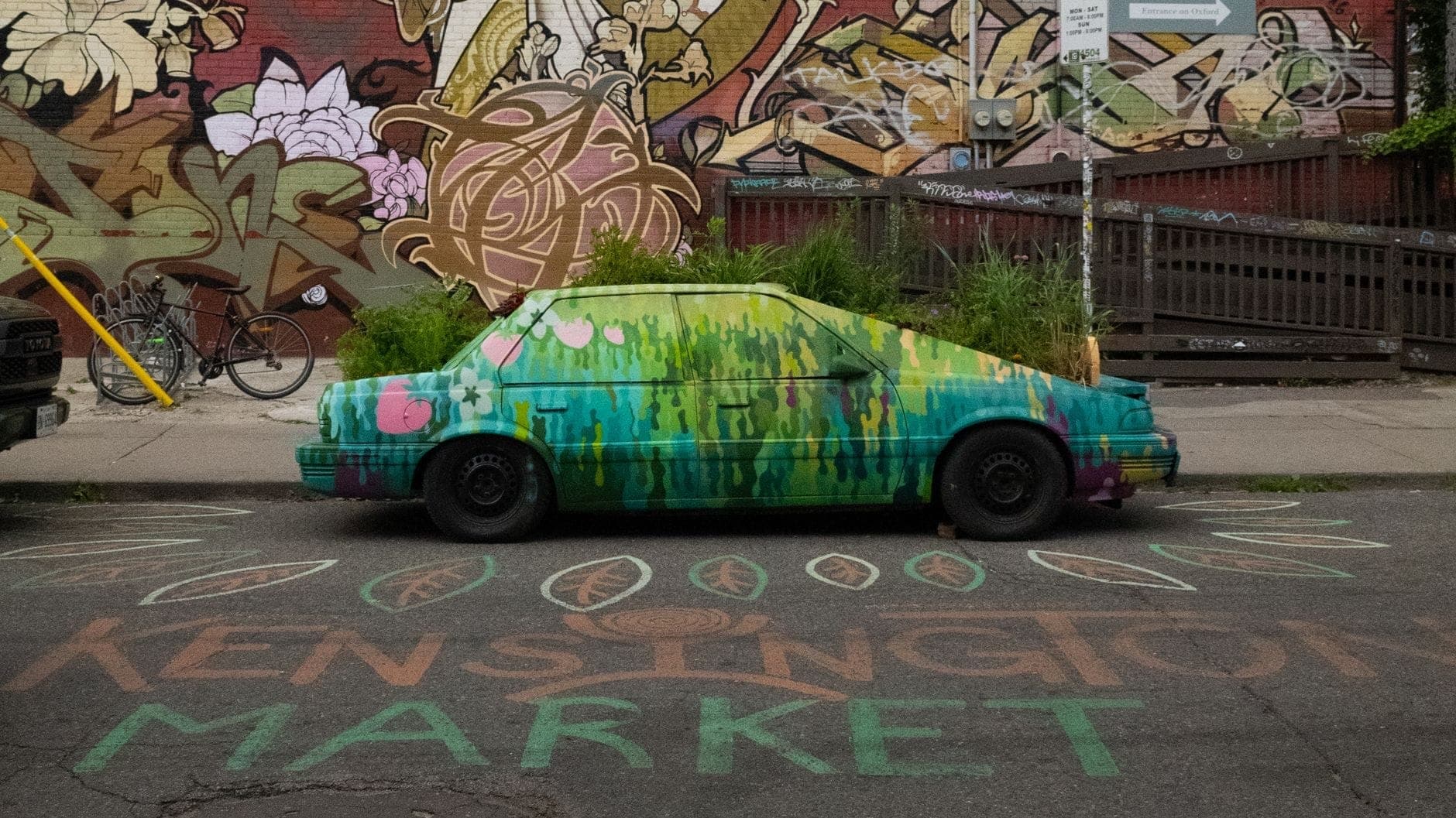 Un'auto dipinta circondata da street art e murales vivaci, con la scritta 'Kensington Market' sull'asfalto, che cattura lo spirito creativo ed eclettico del quartiere.