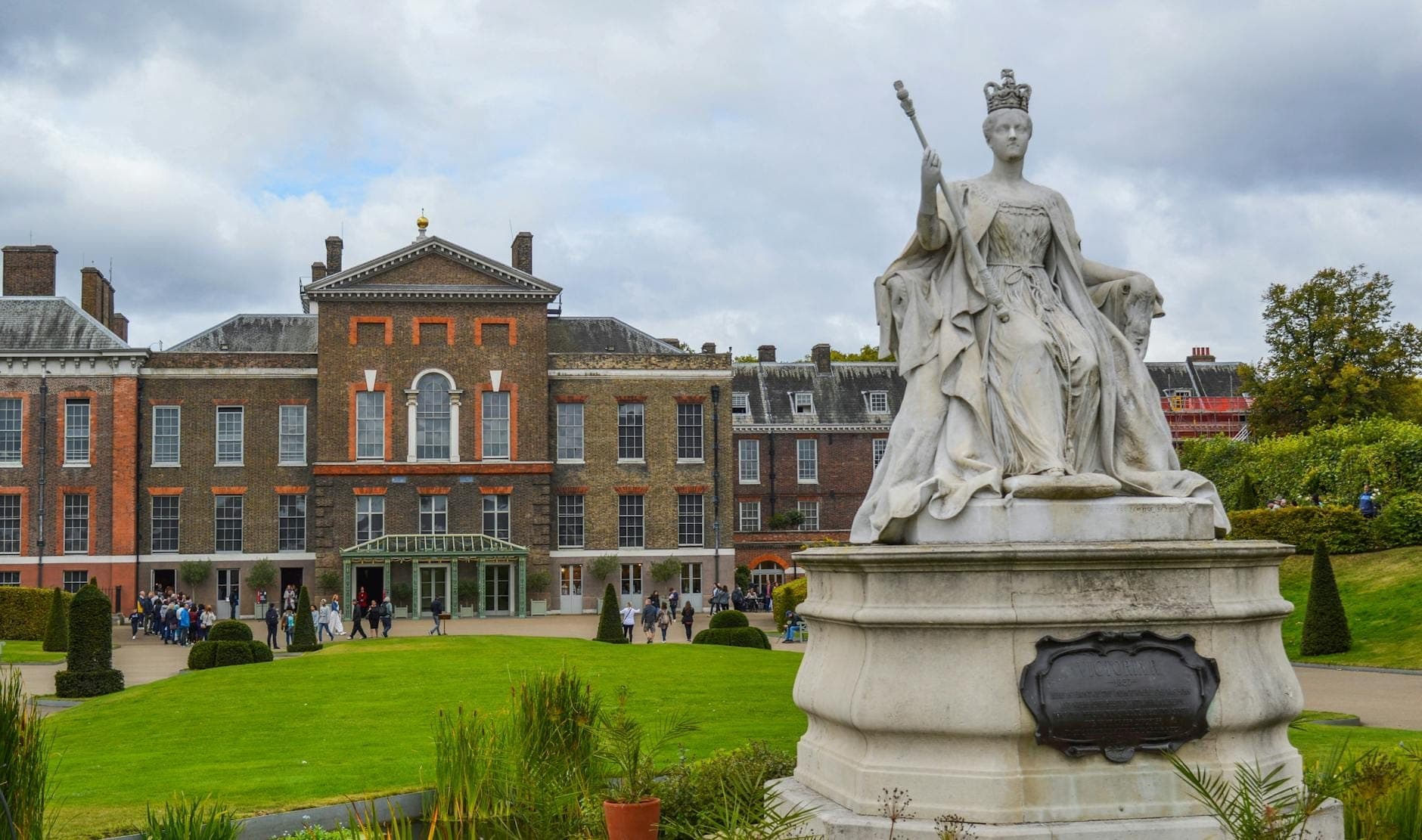 Frontansicht des Kensington Palace mit der Statue von Königin Victoria im Vordergrund und Besuchern in den gepflegten Gartenanlagen.