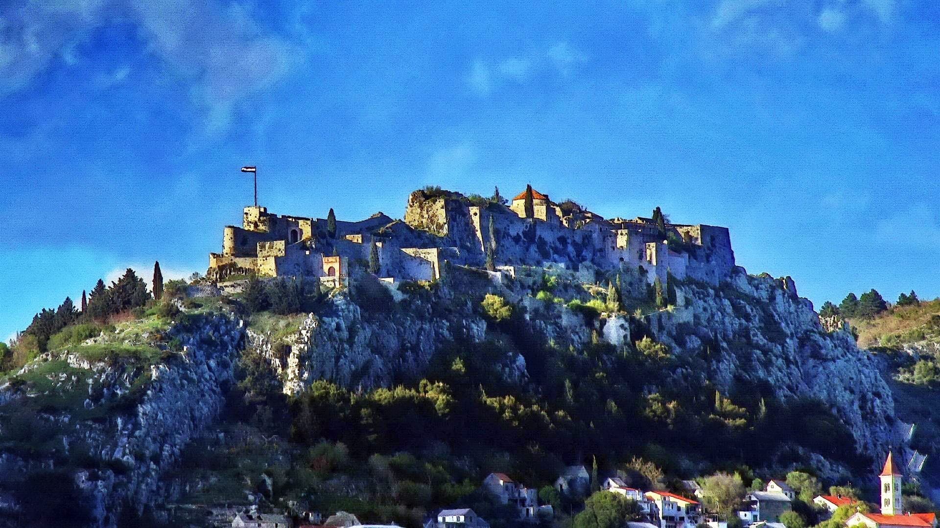 La fortezza di Klis arroccata su una collina rocciosa sopra Split, con mura in pietra e torri illuminate su uno sfondo di cielo azzurro vivido.