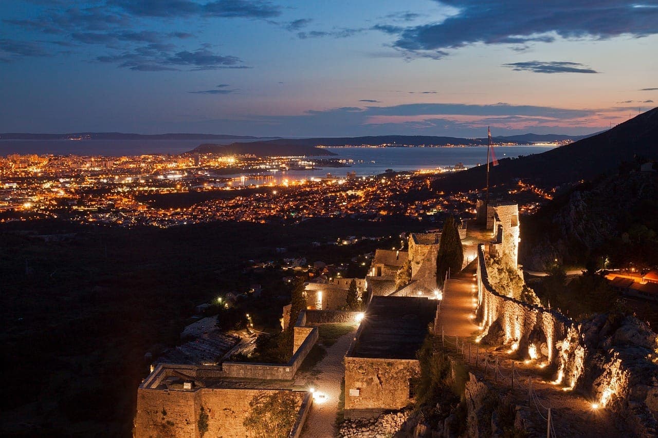 Vista della Fortezza di Klis illuminata al crepuscolo, con le luci scintillanti della città di Split e il Mare Adriatico sullo sfondo.
