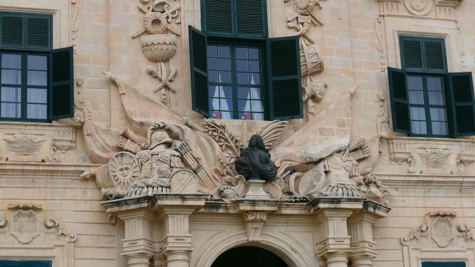 Nahaufnahme einer verzierten Steinfassade mit barocken Schnitzereien und offenen grünen Fensterläden an einem historischen Gebäude in Malta.
