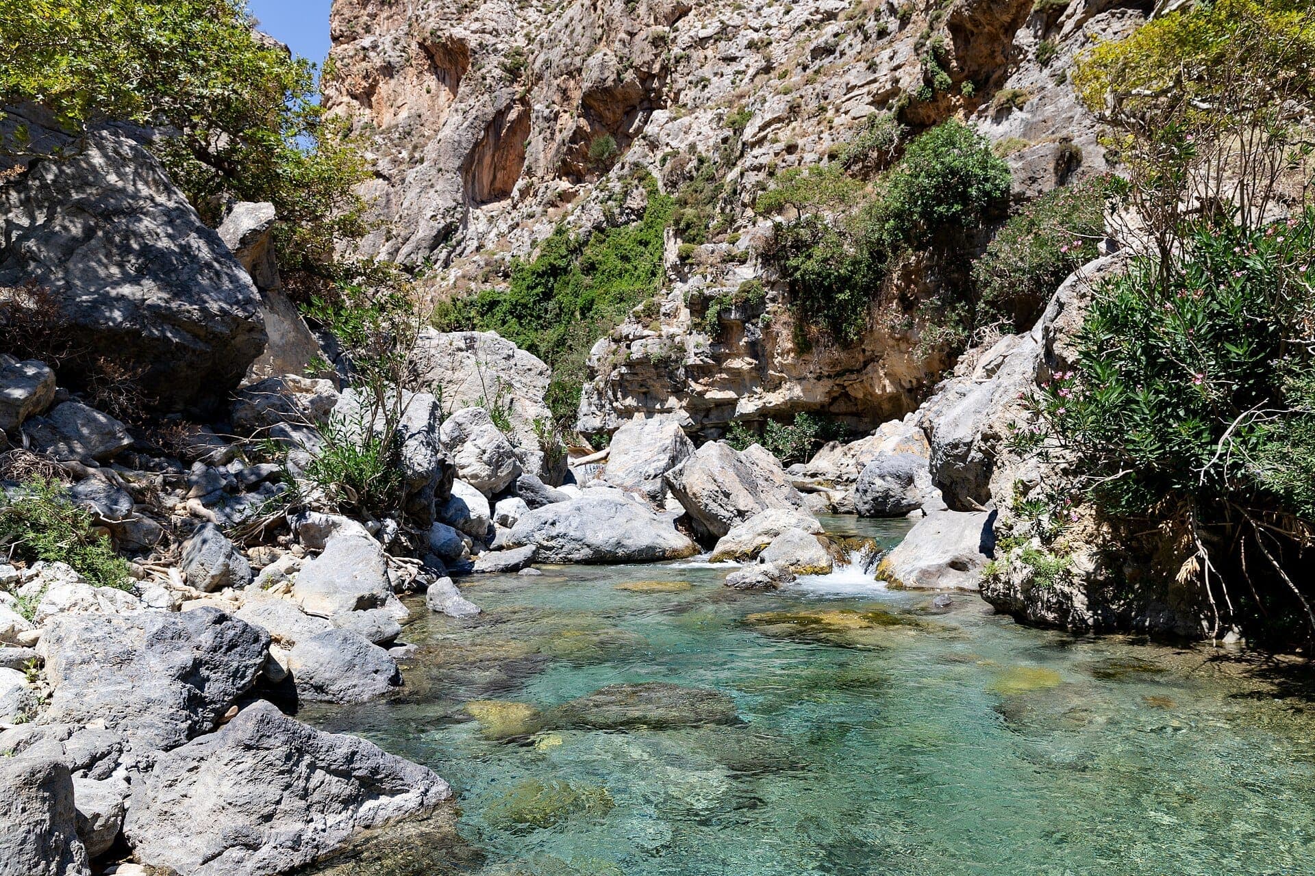 Klarer türkisfarbener Fluss, der durch felsige Kalksteinklippen und grüne Vegetation in der Kourtaliotiko-Schlucht bei hellem Tageslicht fließt.