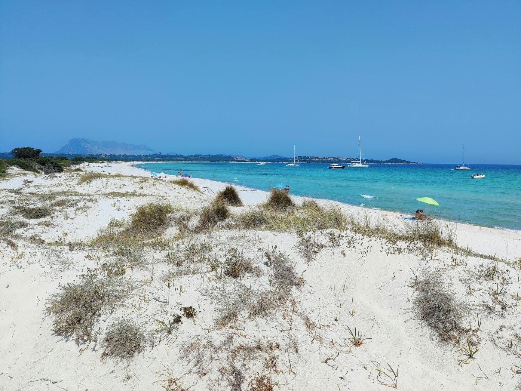 Weitblick über La Cinta Beach mit feinen weißen Sanddünen im Vordergrund und türkisfarbenem Wasser, in dem Segelboote unter einem klaren blauen Himmel vor Anker liegen.