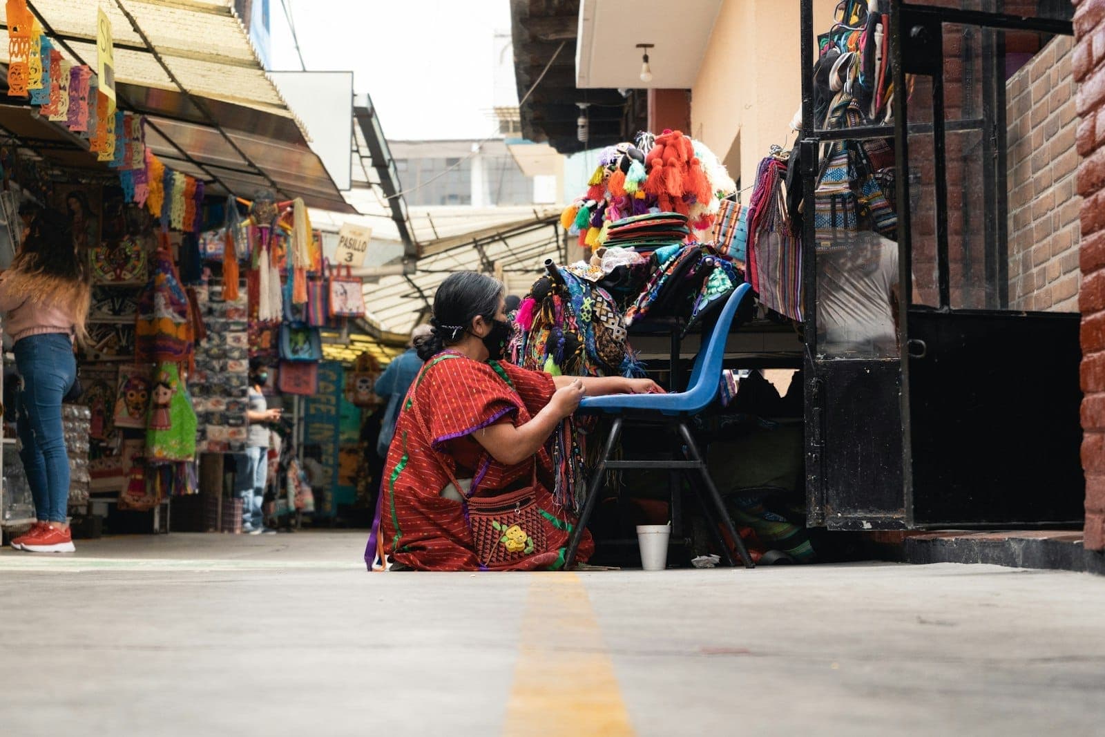 ผู้หญิงในชุดพื้นเมืองเม็กซิกันนั่งอยู่กลางตลาดหัตถกรรม สดใสเต็มไปด้วยงานฝีมือและผ้าทอหลากสีในบรรยากาศร้านค้าสไตล์เปิดโล่ง