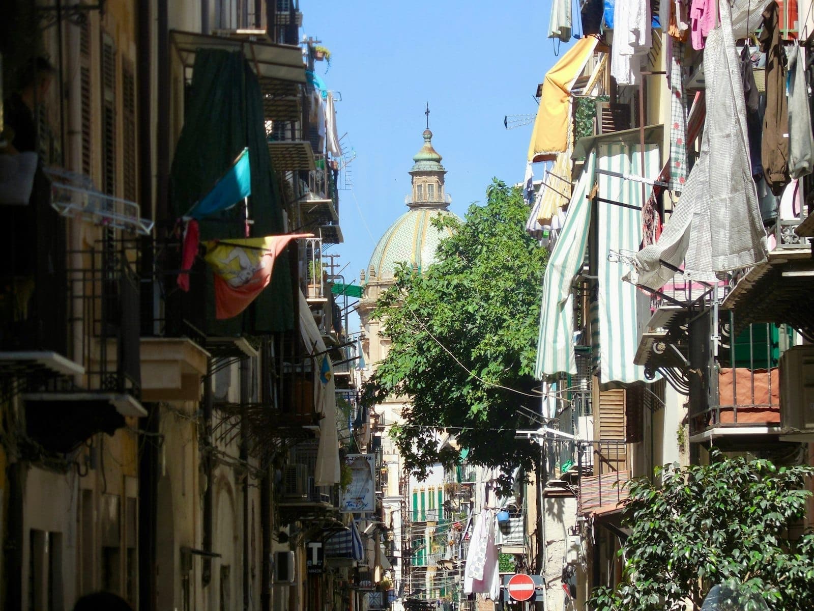 Jalan sempit di La Kalsa, Palermo, diapit bangunan tua, jemuran berwarna-warni yang tergantung dari balkon, dan sebuah gereja berkubah di latar belakang.