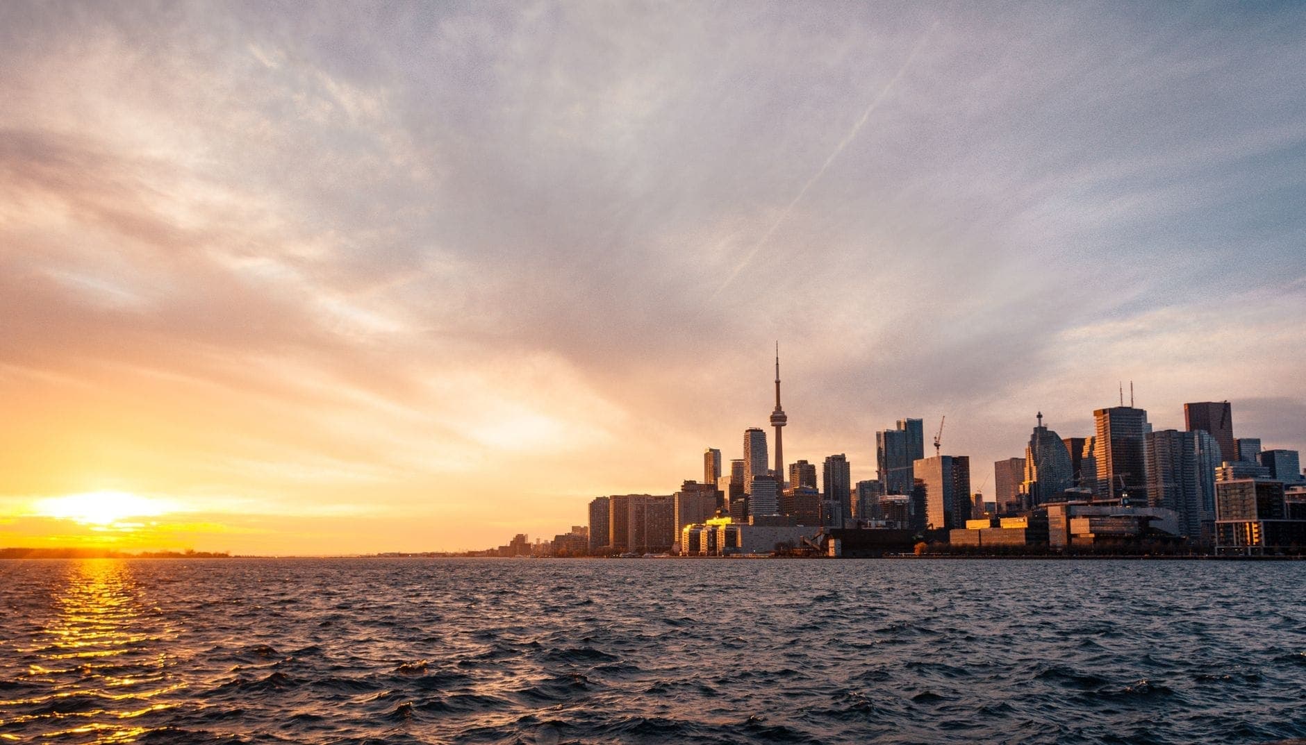 Skyline di Toronto vista dal Lago Ontario al tramonto, con gli edifici sul lungofiume e la CN Tower in silhouette contro un cielo colorato.