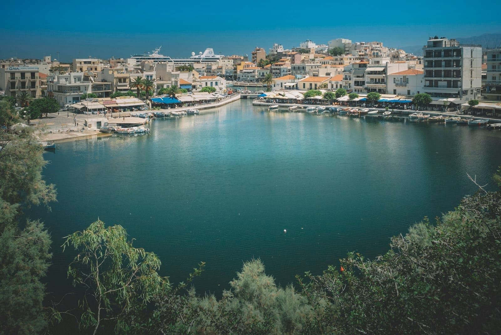 Blick auf den Voulisman-See in Agios Nikolaos mit tiefblauem Wasser, Cafés am Ufer und Stadtgebäuden, die den kraterartigen See unter klarem Himmel umgeben.