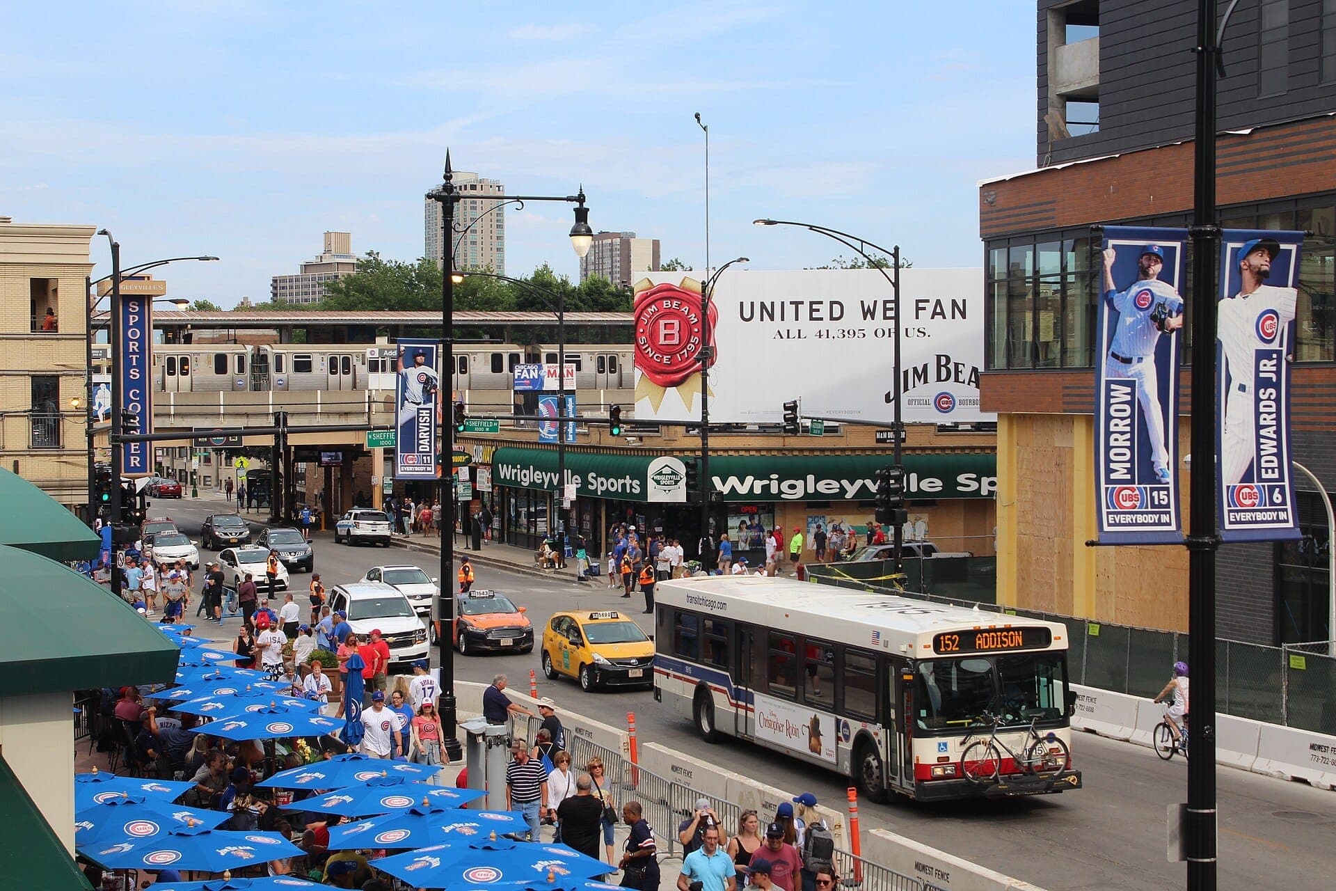 Busy Wrigleyville intersection in Lakeview with crowds, CTA bus, taxis, and elevated train beside restaurants and sports bars on a sunny day.