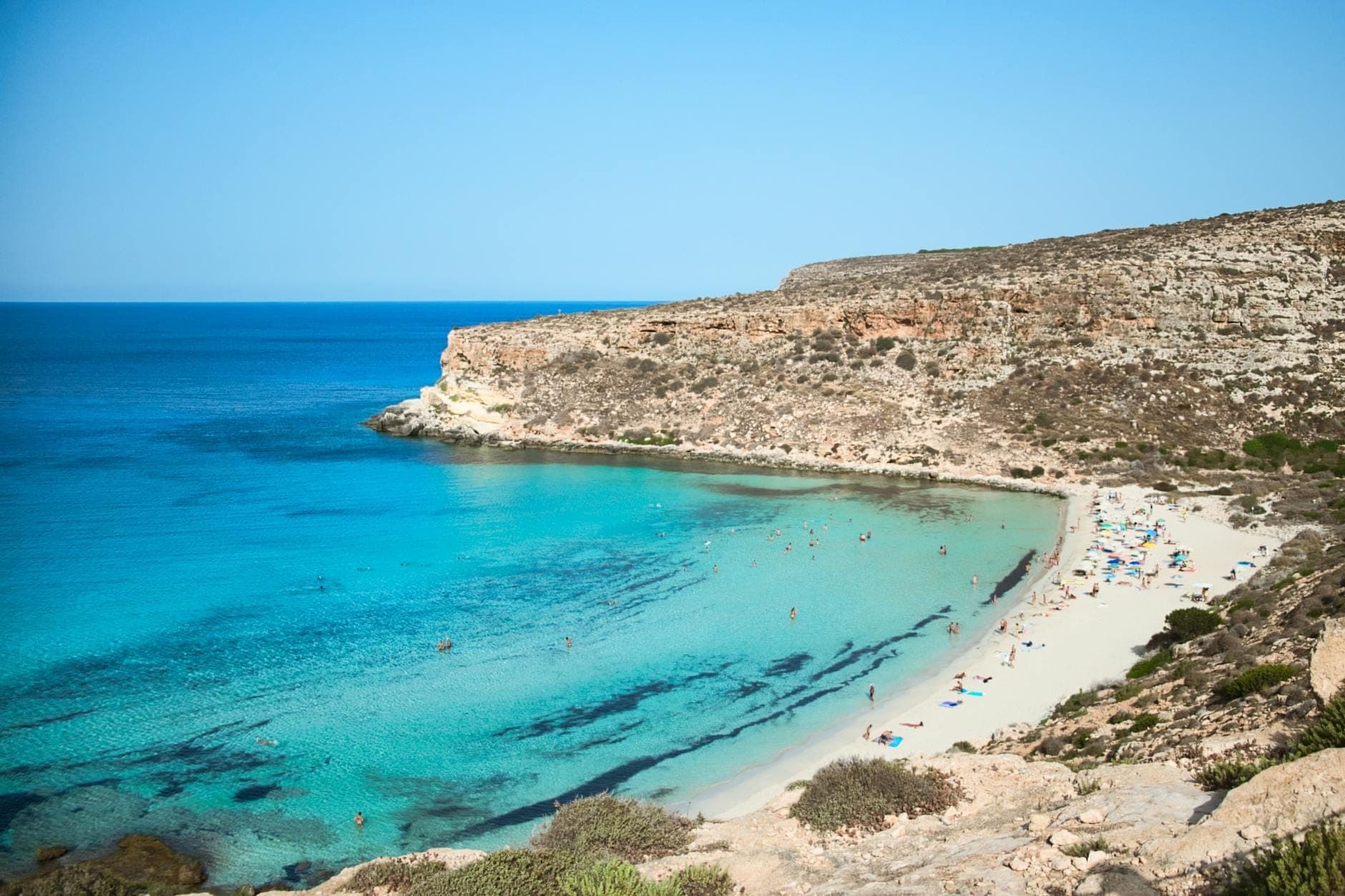 Pemandangan luas Spiaggia dei Conigli dengan air toska yang cerah, pantai berpasir putih, pengunjung yang berjemur, dan tebing berbatu di bawah langit biru jernih di Lampedusa.