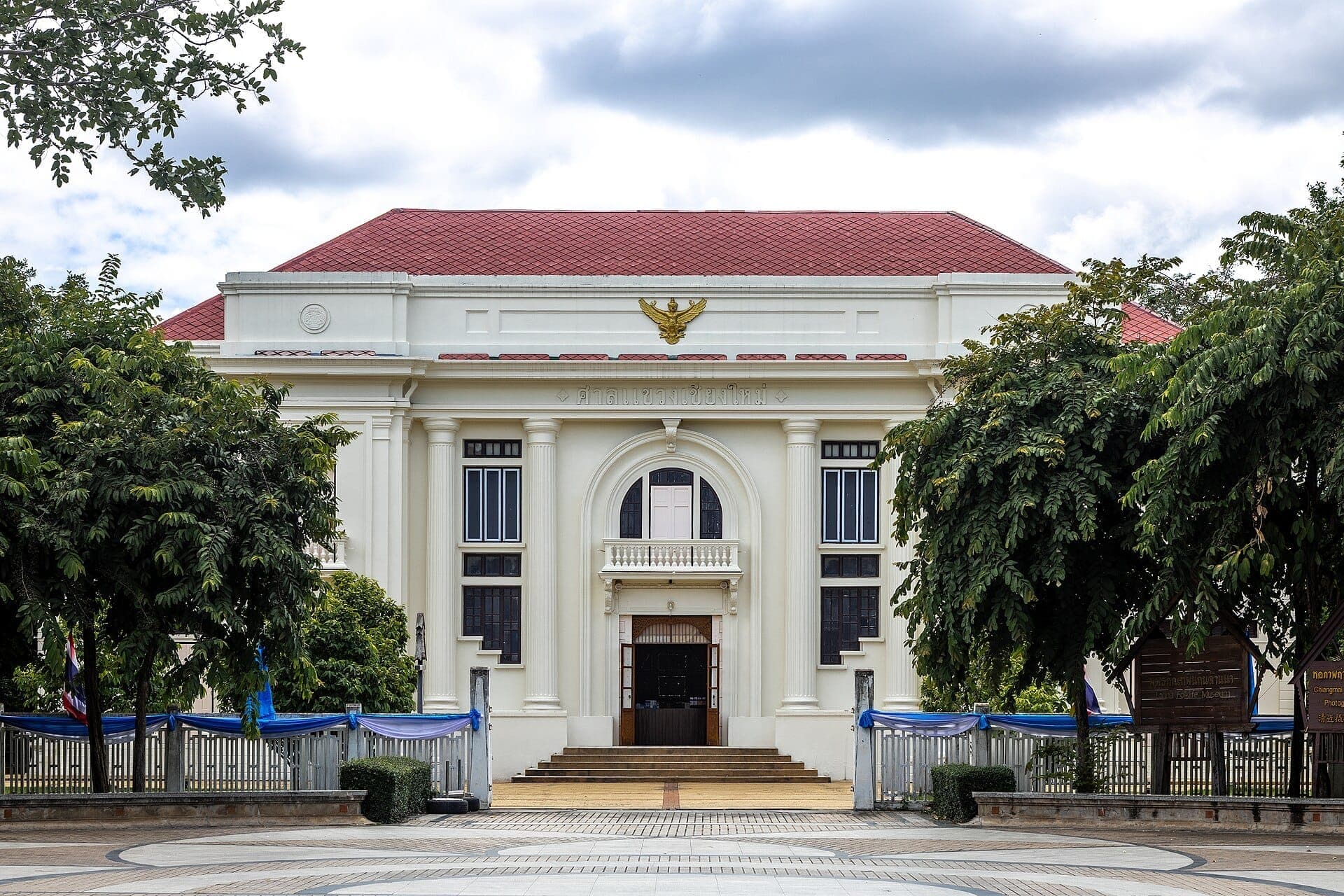Vorderansicht des Lanna Folklife Museum in Chiang Mai mit einem weißen Gebäude aus der Kolonialzeit, rotem Dach und üppigen grünen Bäumen.