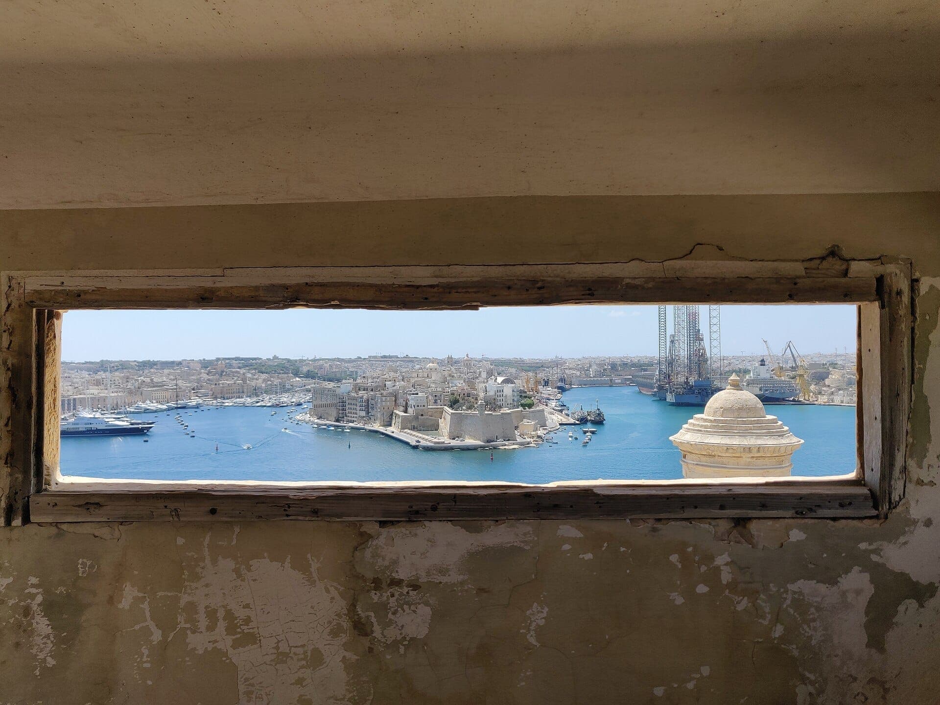 Blick auf den Grand Harbour von Valletta und die Stadtsilhouette durch ein verwittertes Fenster in den Lascaris War Rooms, mit historischen Steinmauern und blauem Meer.