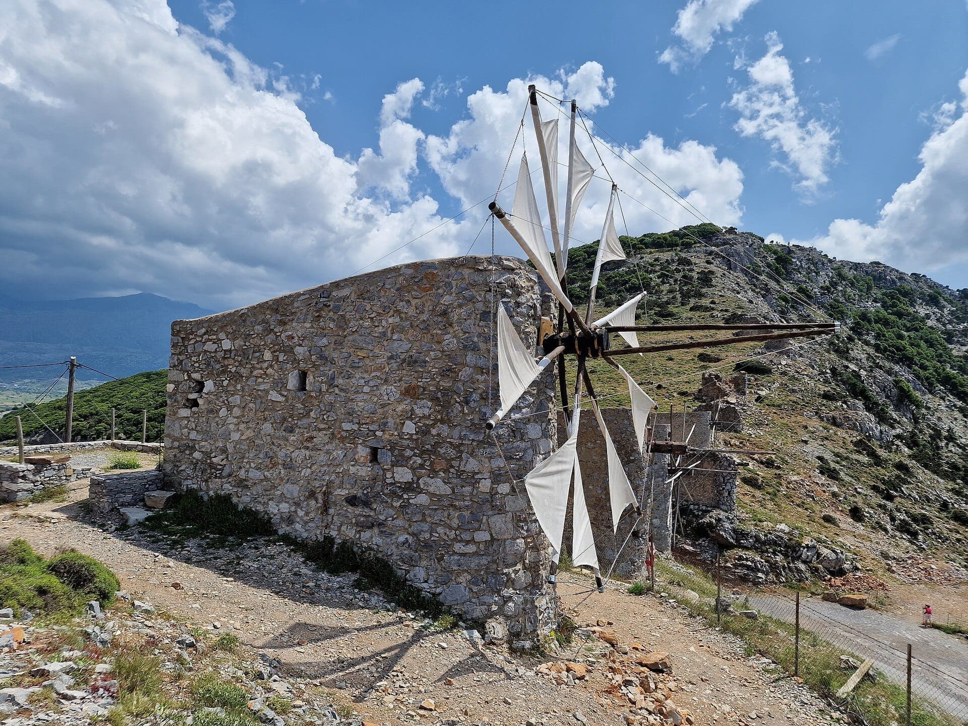 Steinwindmühle mit weißen Segeln aus Leinwand auf dem Lassithi-Plateau, im Hintergrund zerklüftete Hügel und ein heller, teils bewölkter Himmel auf Kreta.