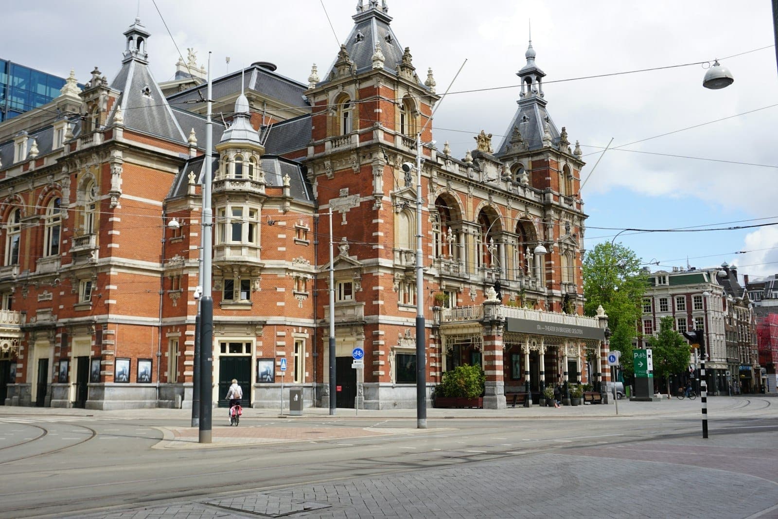 Stadsschouwburg-Theater am Leidseplein in Amsterdam mit seiner kunstvollen Backsteinfassade, Straßenbahnschienen und einem Radfahrer an einem bewölkten Tag.