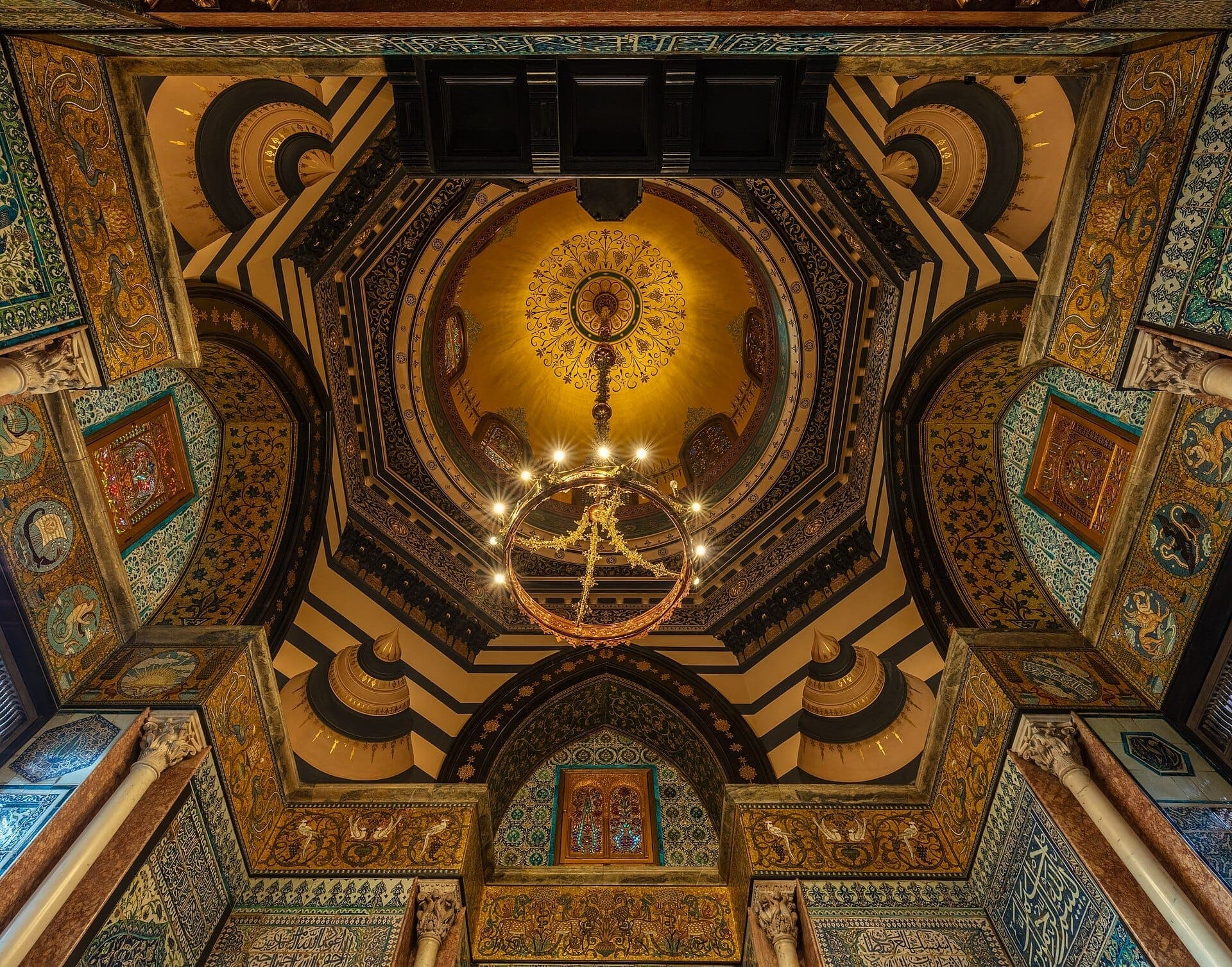 Zdobiony kopułowy sufit Arab Hall w Leighton House Museum, z pozłacanymi detalami, łukami w paski, płytkami Iznik i centralnym żyrandolem.
