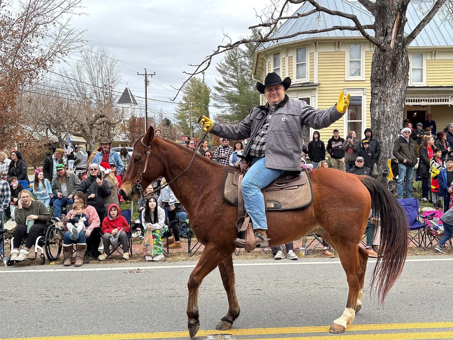 A cowboy on horseback waves to a crowd during a Leiper's Fork Village parade, with historic homes and winter trees in the background.