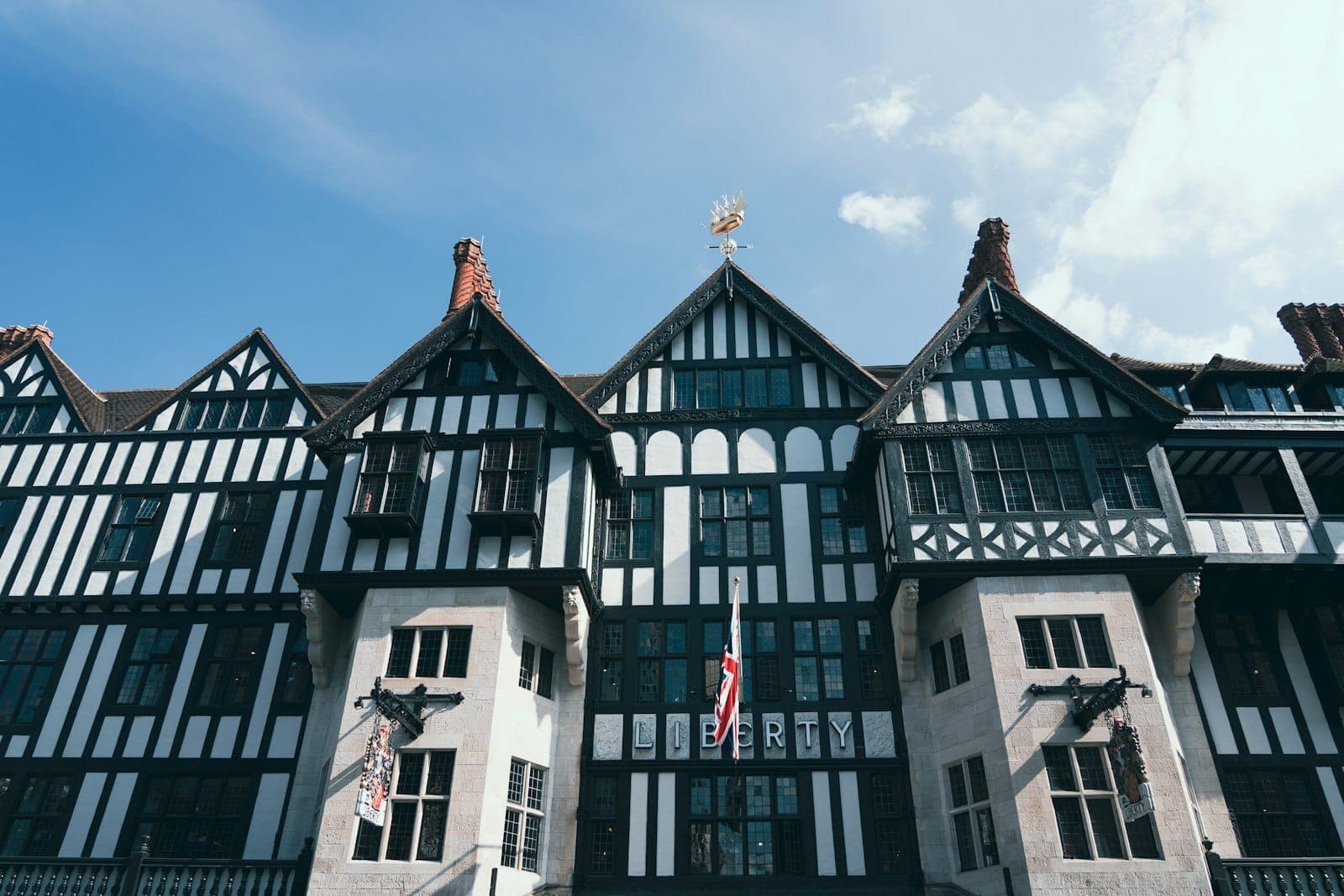 Die schwarz-weiße Fachwerkfassade des Kaufhauses Liberty London mit Giebeldächern, Bleiglasfenstern und der Union Jack an einem sonnigen Tag.