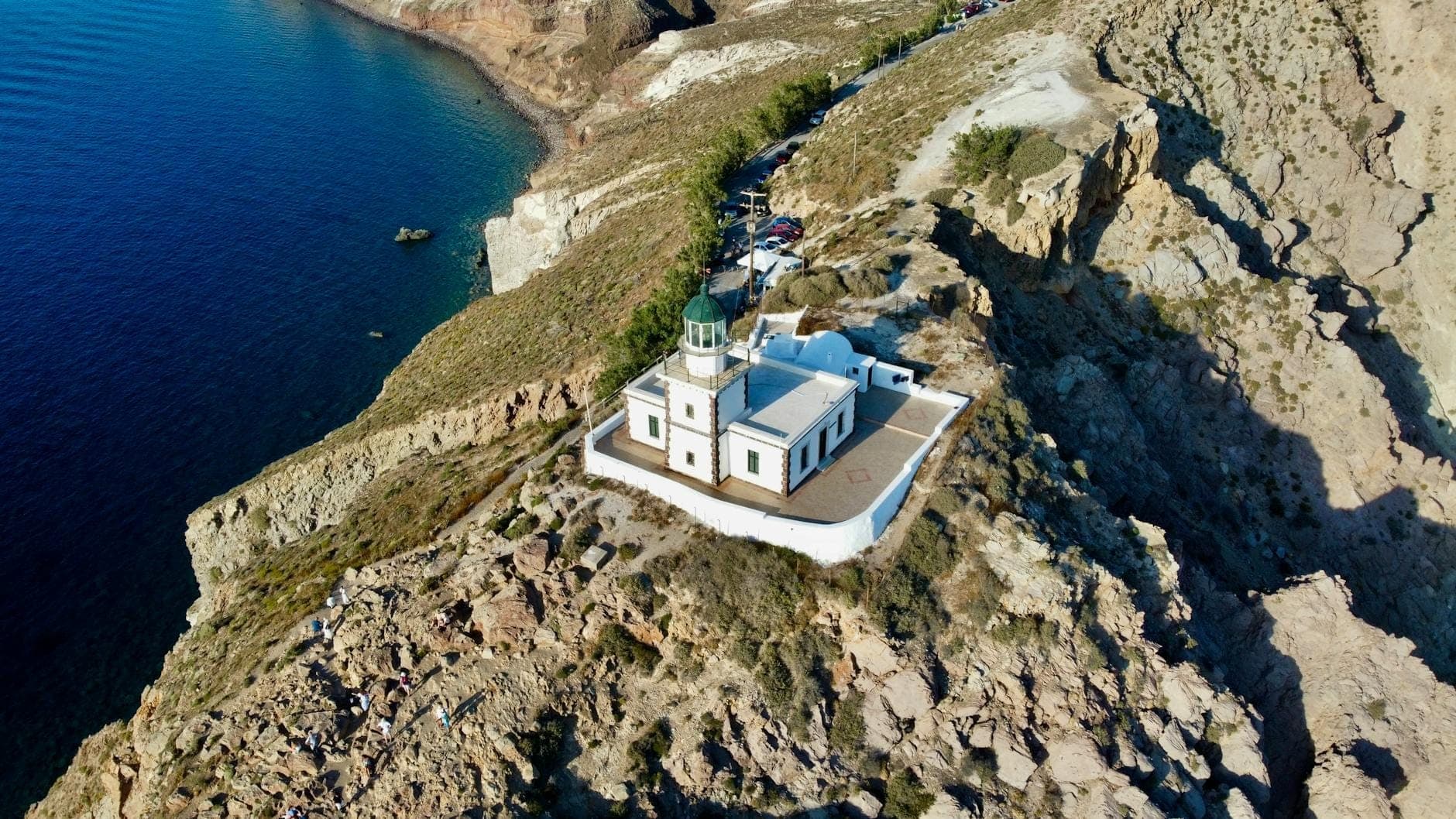 Luftaufnahme des Leuchtturms Akrotiri auf den zerklüfteten Klippen an der Südwestspitze Santorinis, mit tiefblauer Ägäis, dramatischer Küstenlinie und klarem Himmel.
