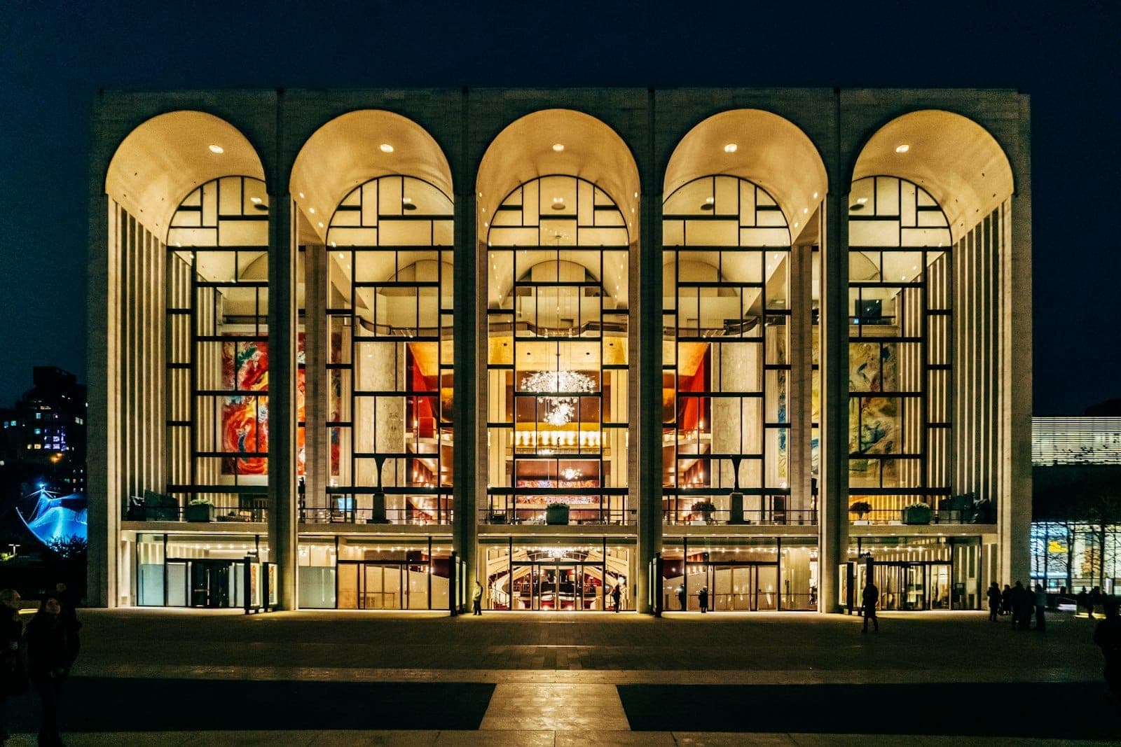 วิวภายนอกของ Lincoln Center for the Performing Arts ในยามค่ำคืน พร้อมซุ้มโค้งที่สว่างไสวและหน้าต่างกระจกขนาดใหญ่หันหน้าสู่ลานกว้าง