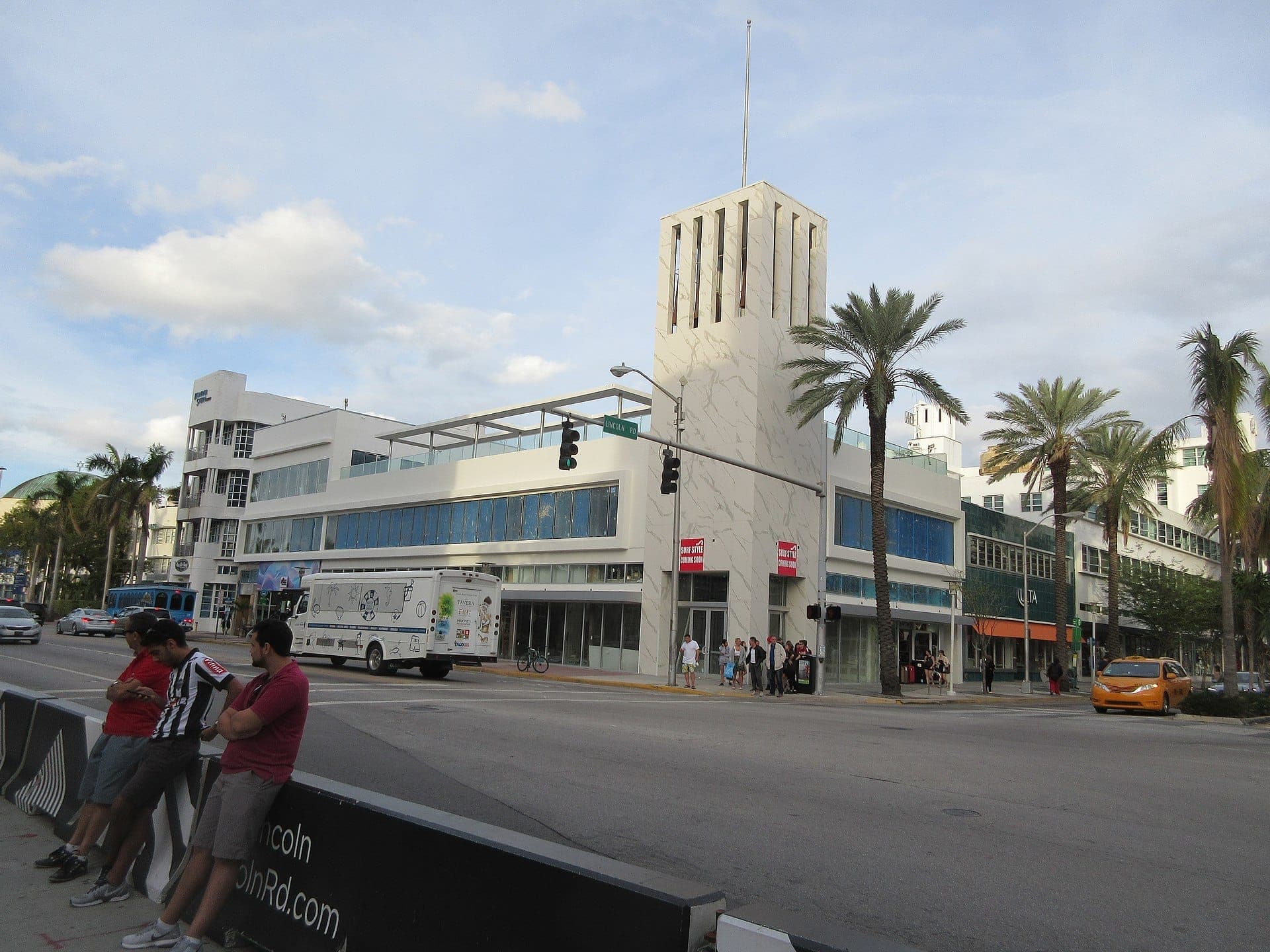 Edificios Art Déco, palmeras y peatones bordean el vibrante Lincoln Road Mall en South Beach bajo un cielo parcialmente nublado.