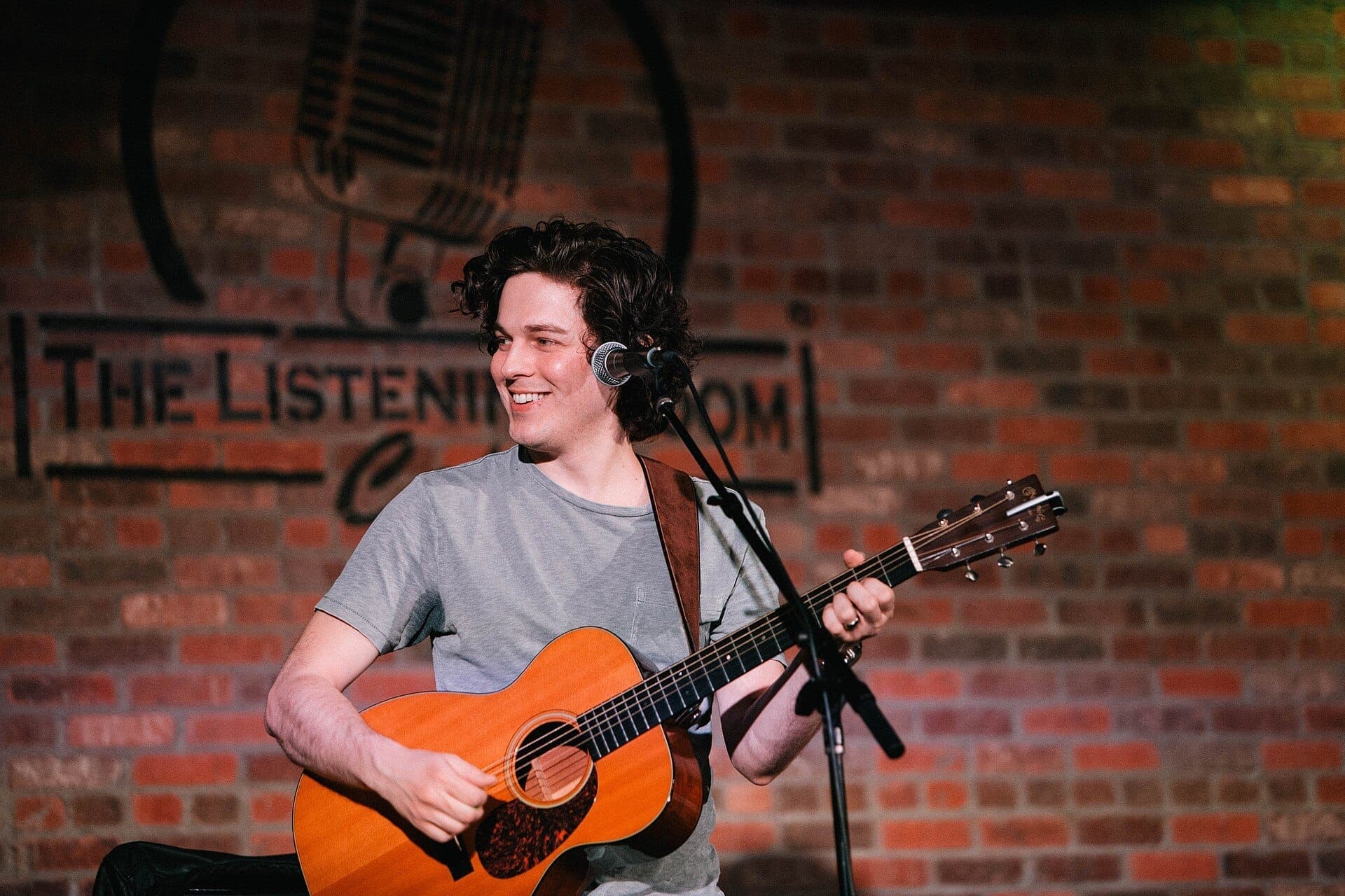 A smiling Ryan Harris Brown plays acoustic guitar on stage at The Listening Room Cafe with a brick wall and logo in the background.