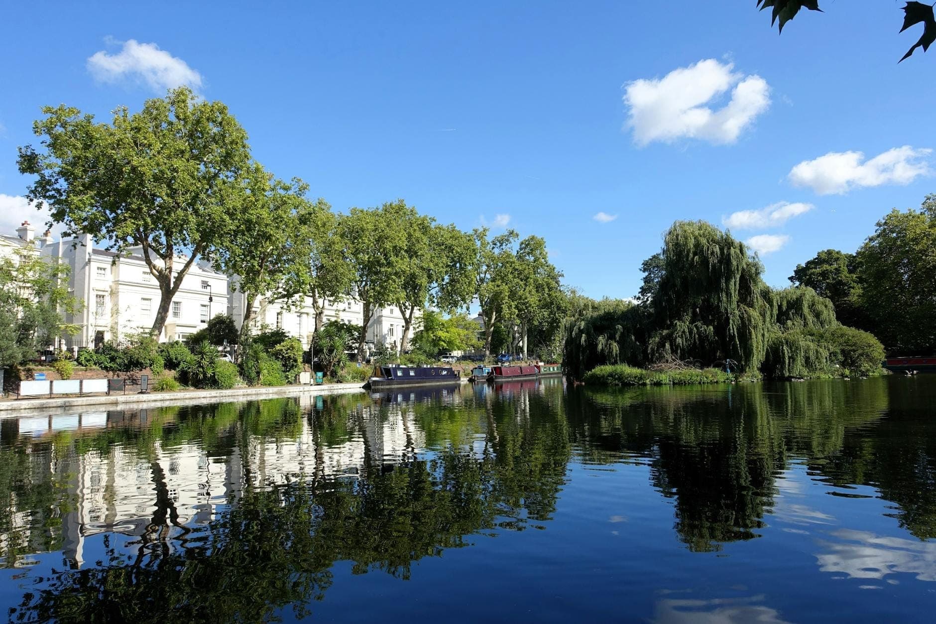 Panoramiczny widok na spokojne wody kanału w Little Venice w Londynie — wąskie łodzie, drzewa w zieleni i eleganckie białe kamienice w stylu regencji odbijają się w wodzie w słoneczny dzień.