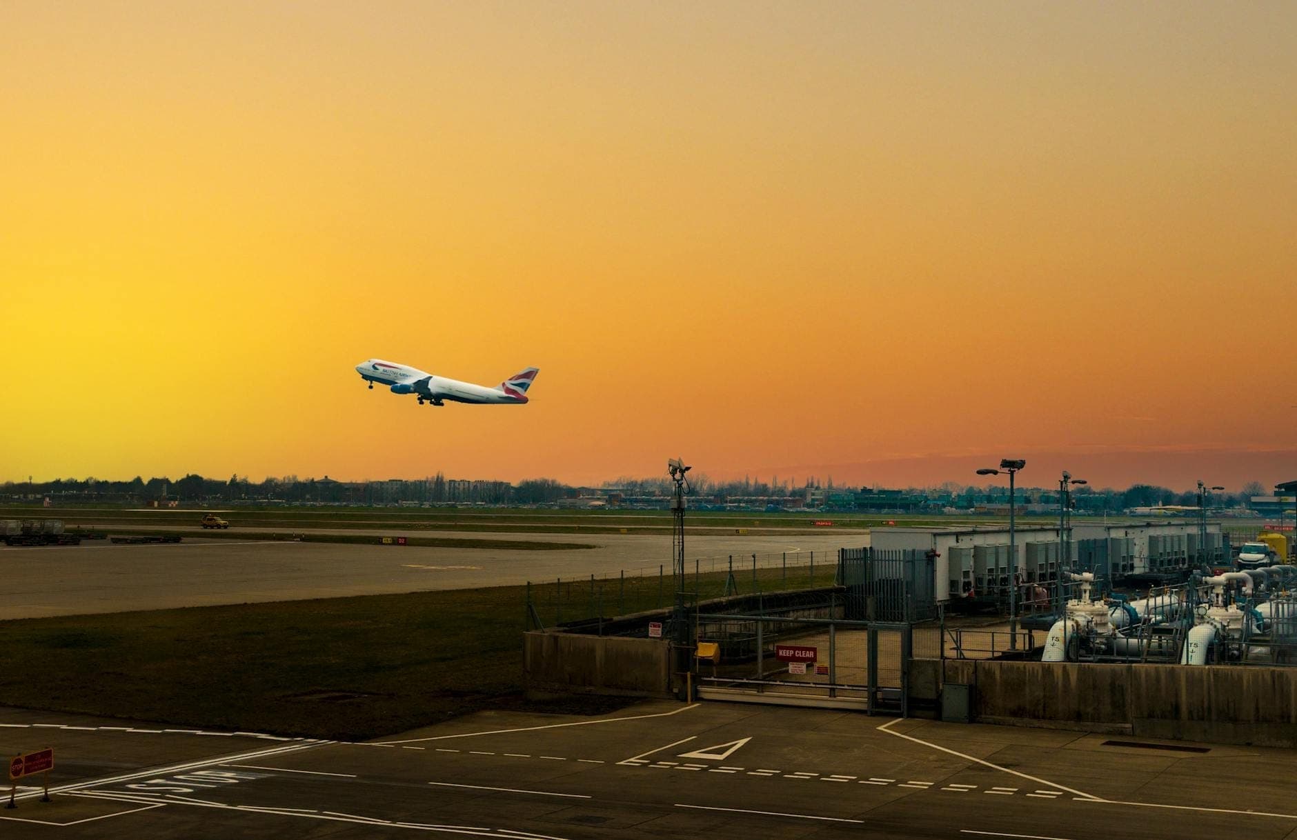 Un aereo di British Airways in decollo da un aeroporto londinese al tramonto, con le infrastrutture aeroportuali, la pista e un ampio cielo sullo sfondo.