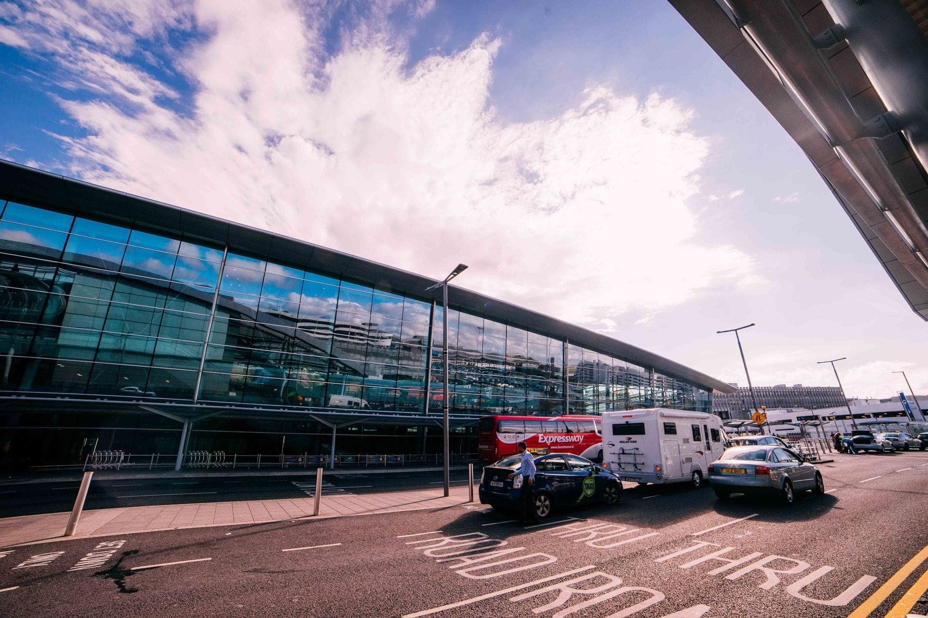 Vista esterna di un moderno terminal in vetro in un aeroporto affollato con veicoli, taxi e passeggeri davanti.
