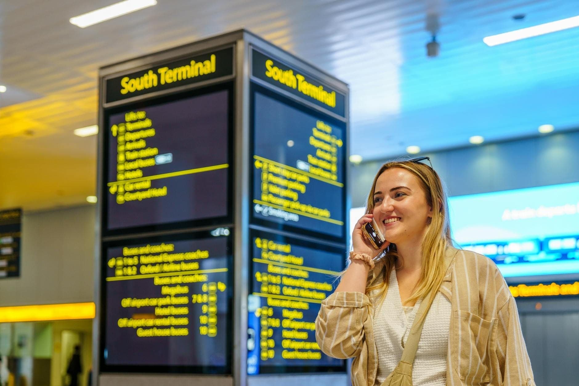 Viaggiatore sorridente al telefono nel Terminal Sud dell'aeroporto di Gatwick, con i tabelloni delle informazioni sui voli visibili sullo sfondo.