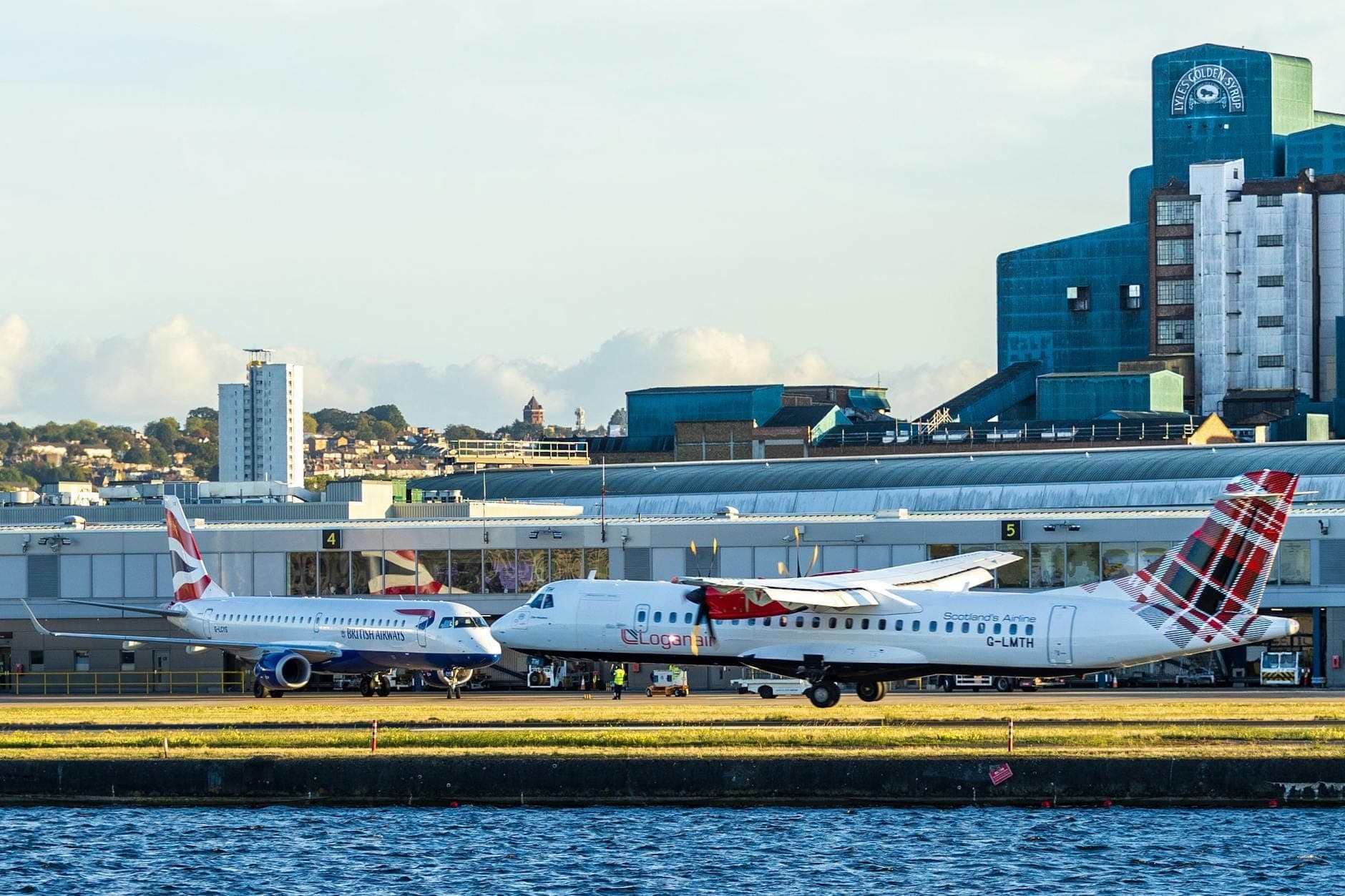 Due aerei commerciali parcheggiati al London City Airport, con edifici moderni e il terminal sullo sfondo, accanto all'acqua.