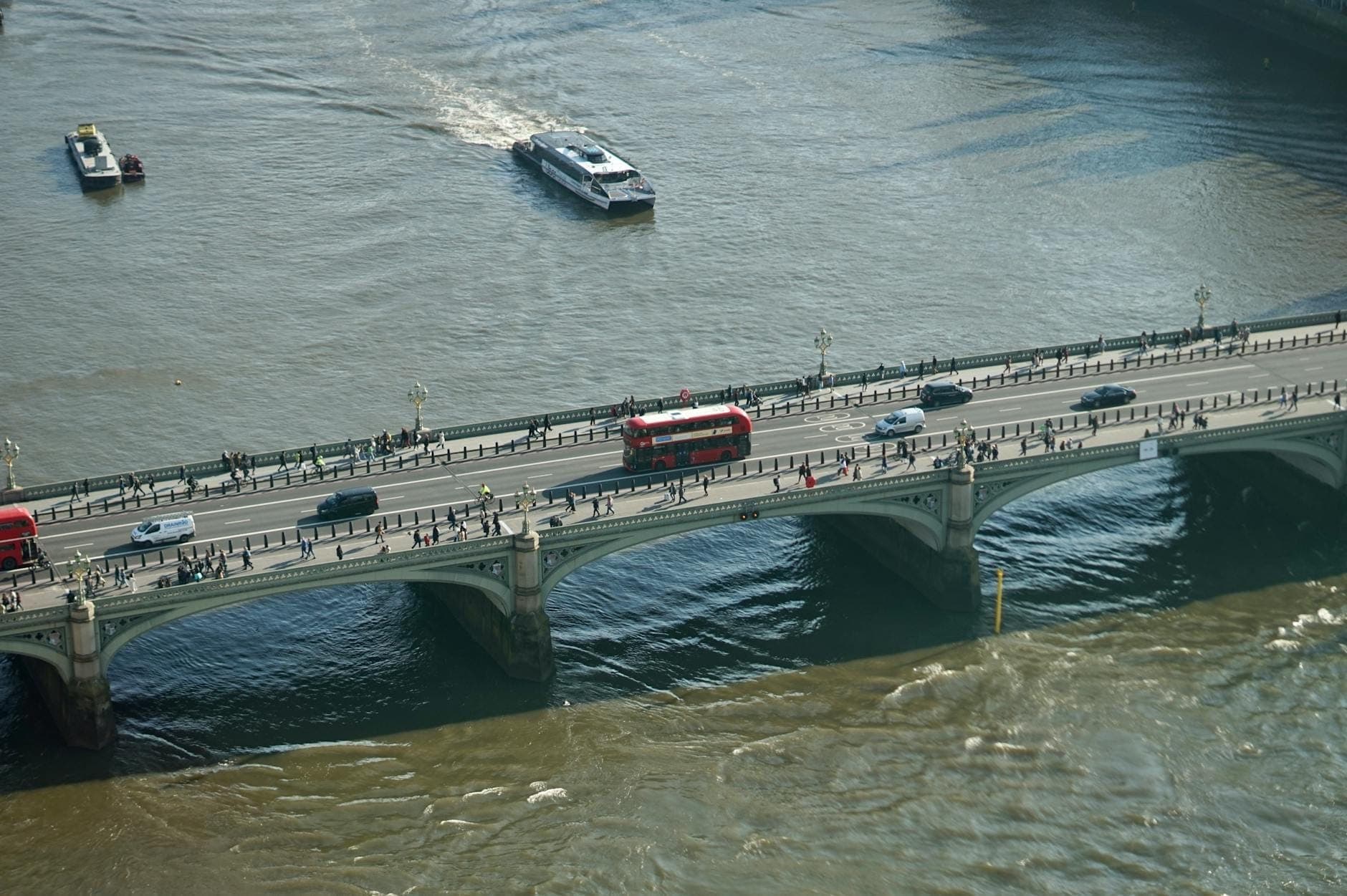 Vista aerea di London Bridge che attraversa il Tamigi con un autobus rosso a due piani, auto e pedoni, barche sul fiume e palazzi sullo sfondo.