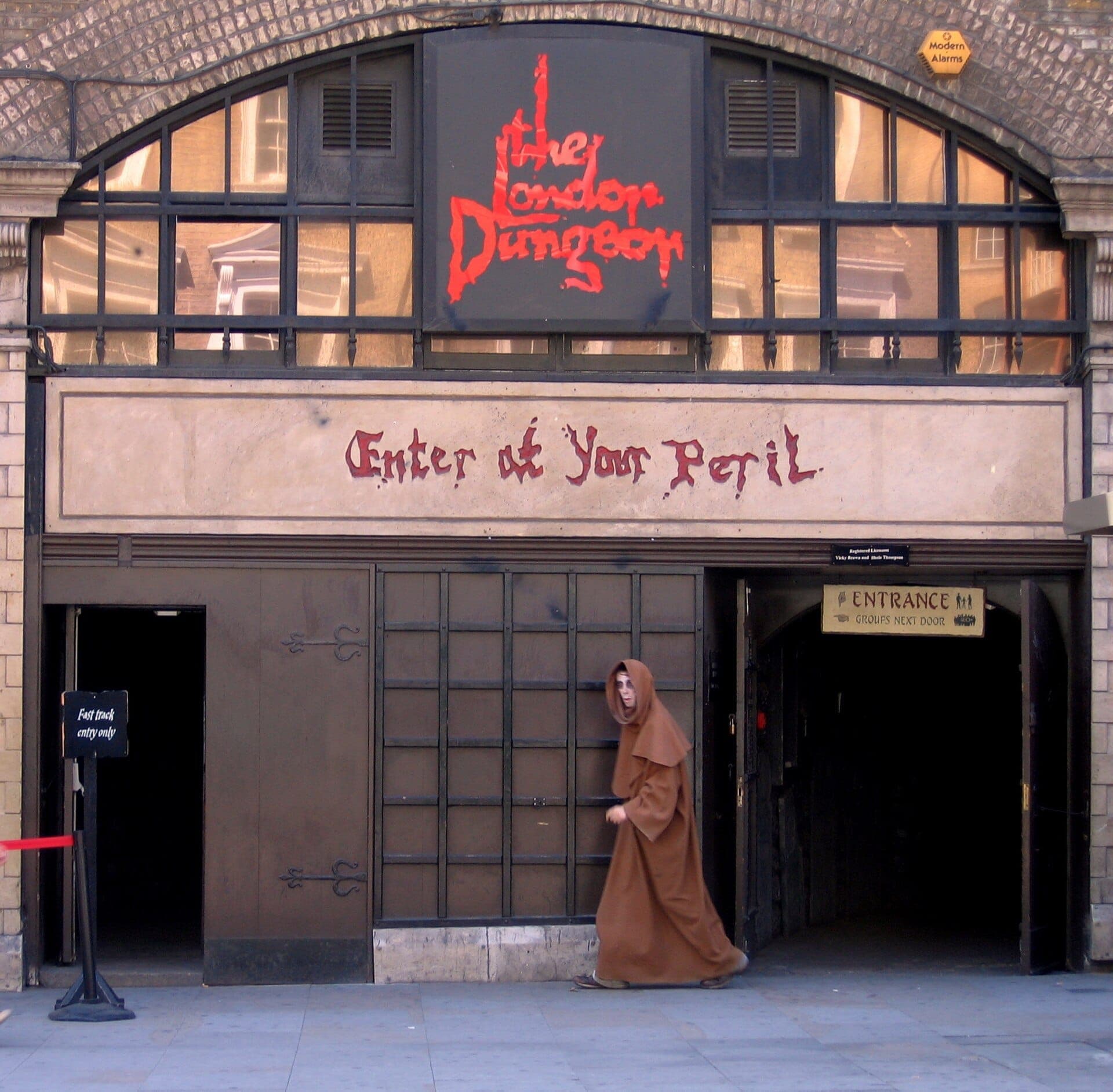 Entrada a The London Dungeon con letreros góticos, ventanas arqueadas y un actor disfrazado con una capa marrón caminando afuera.